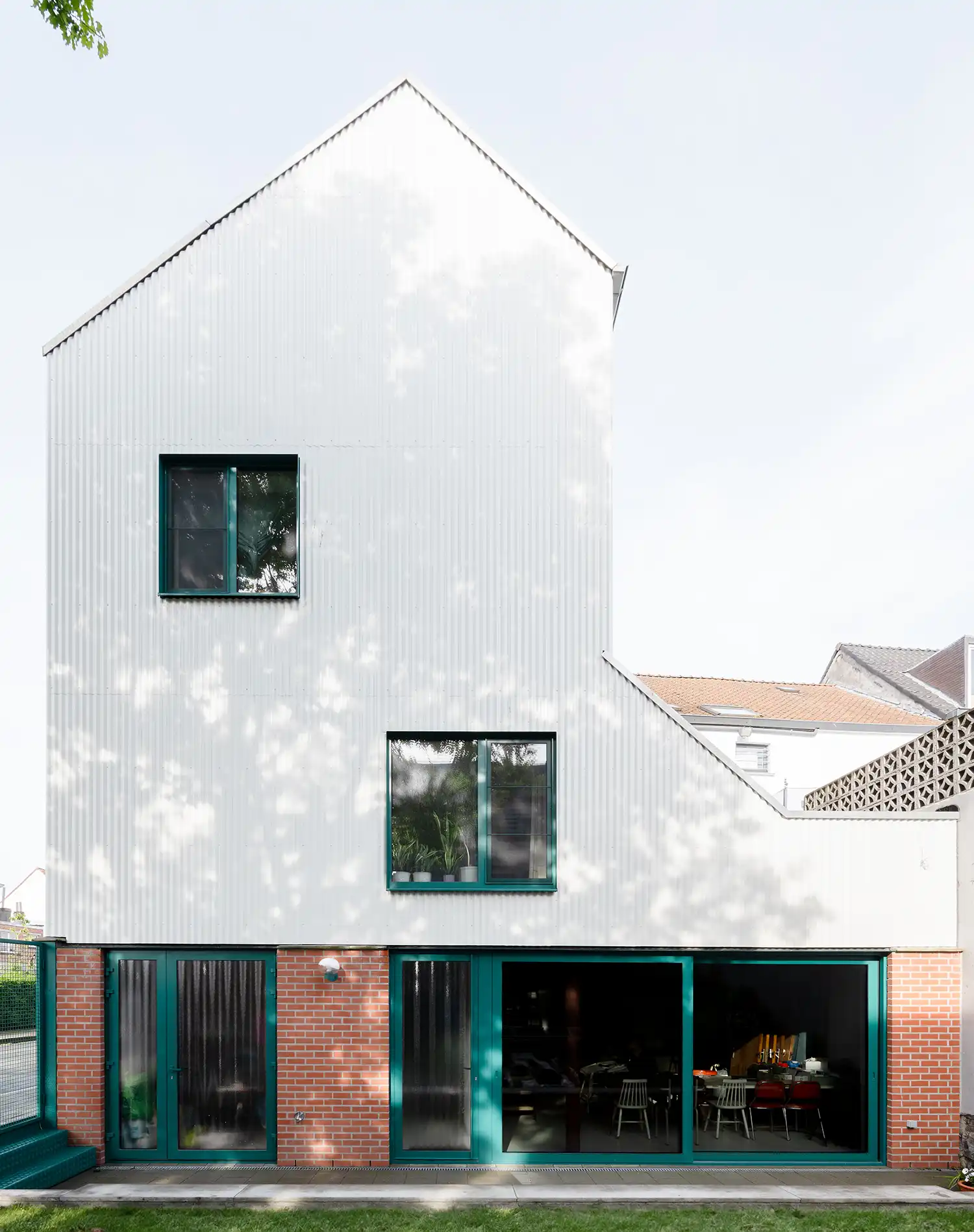 A full view of the back of the house, showing the white corrugated metal upper floors, red brick ground floor with large green-framed glass doors, and a small lawn.