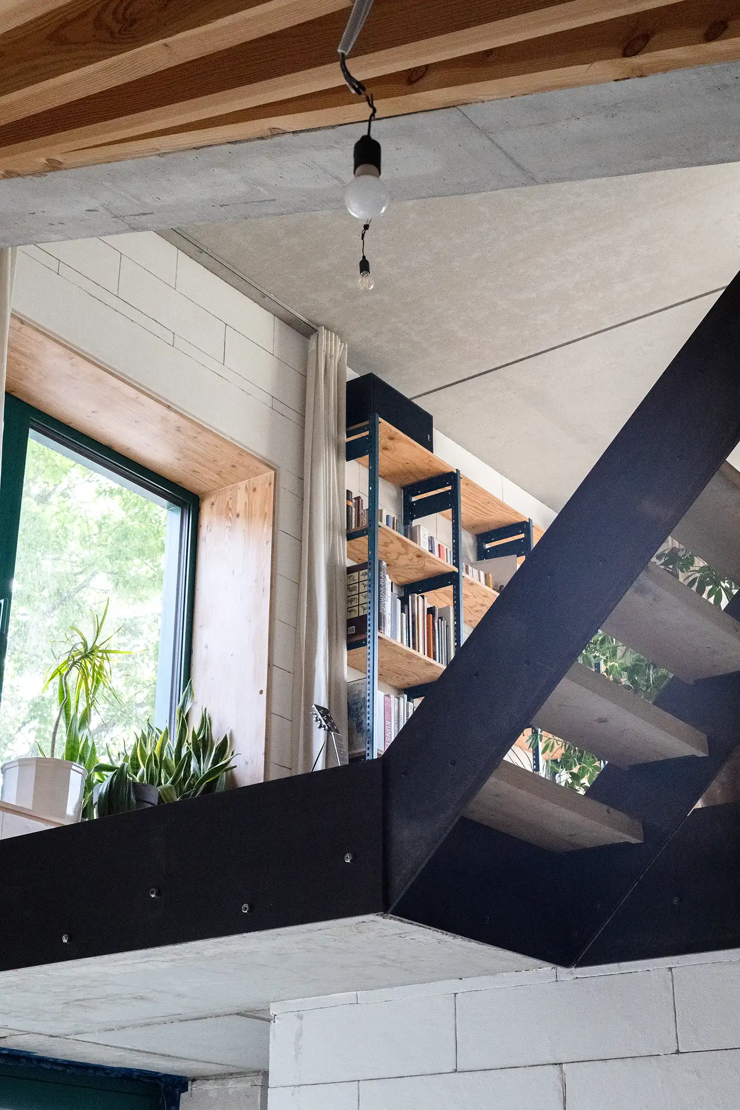 View looking up from the ground floor towards a mezzanine level, with a large window, exposed wooden beams, concrete ceiling, and the underside of a metal staircase.