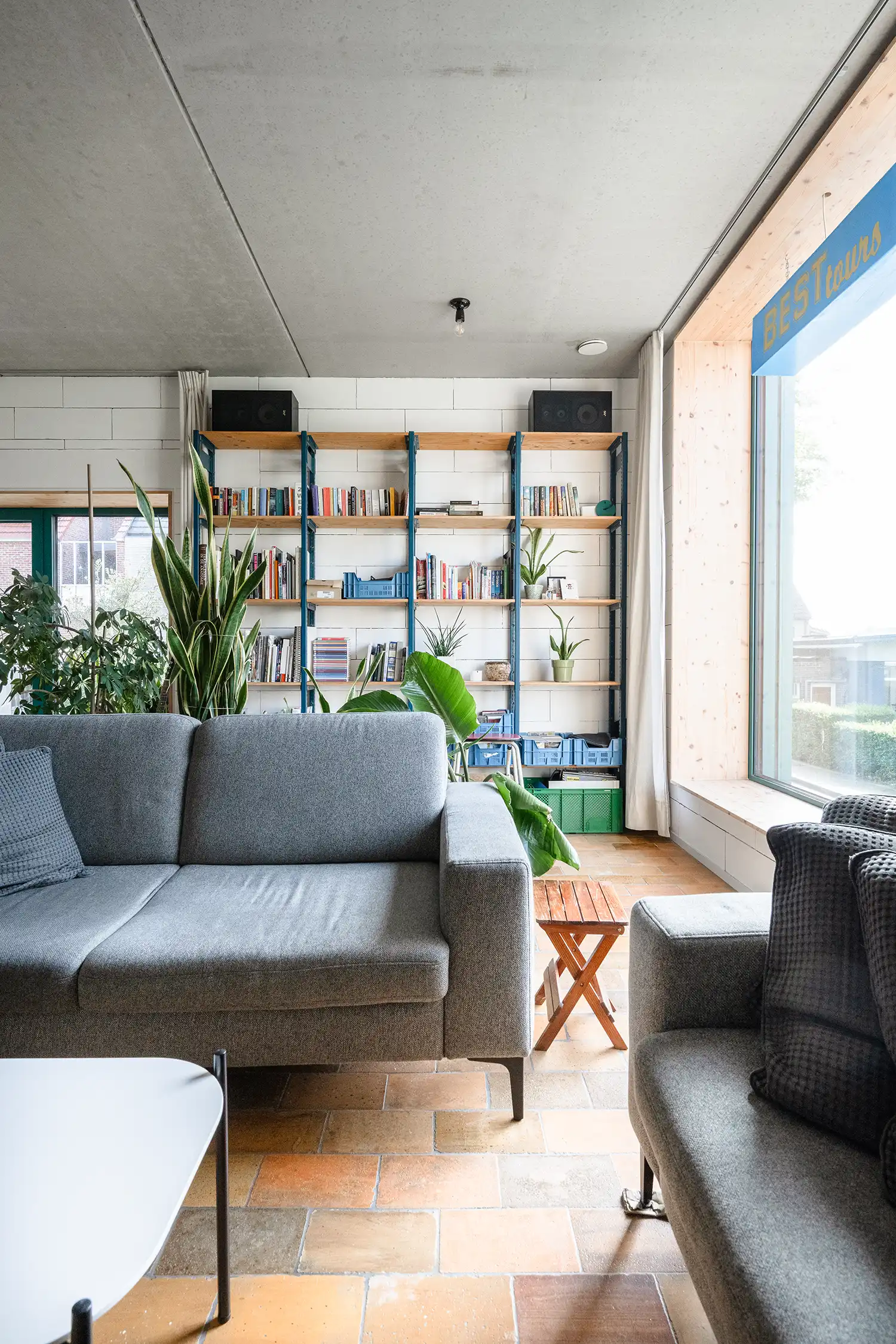 A living room corner with a large grey sofa, a tall wooden bookshelf filled with books and plants, a concrete ceiling, and a large window with green frames.