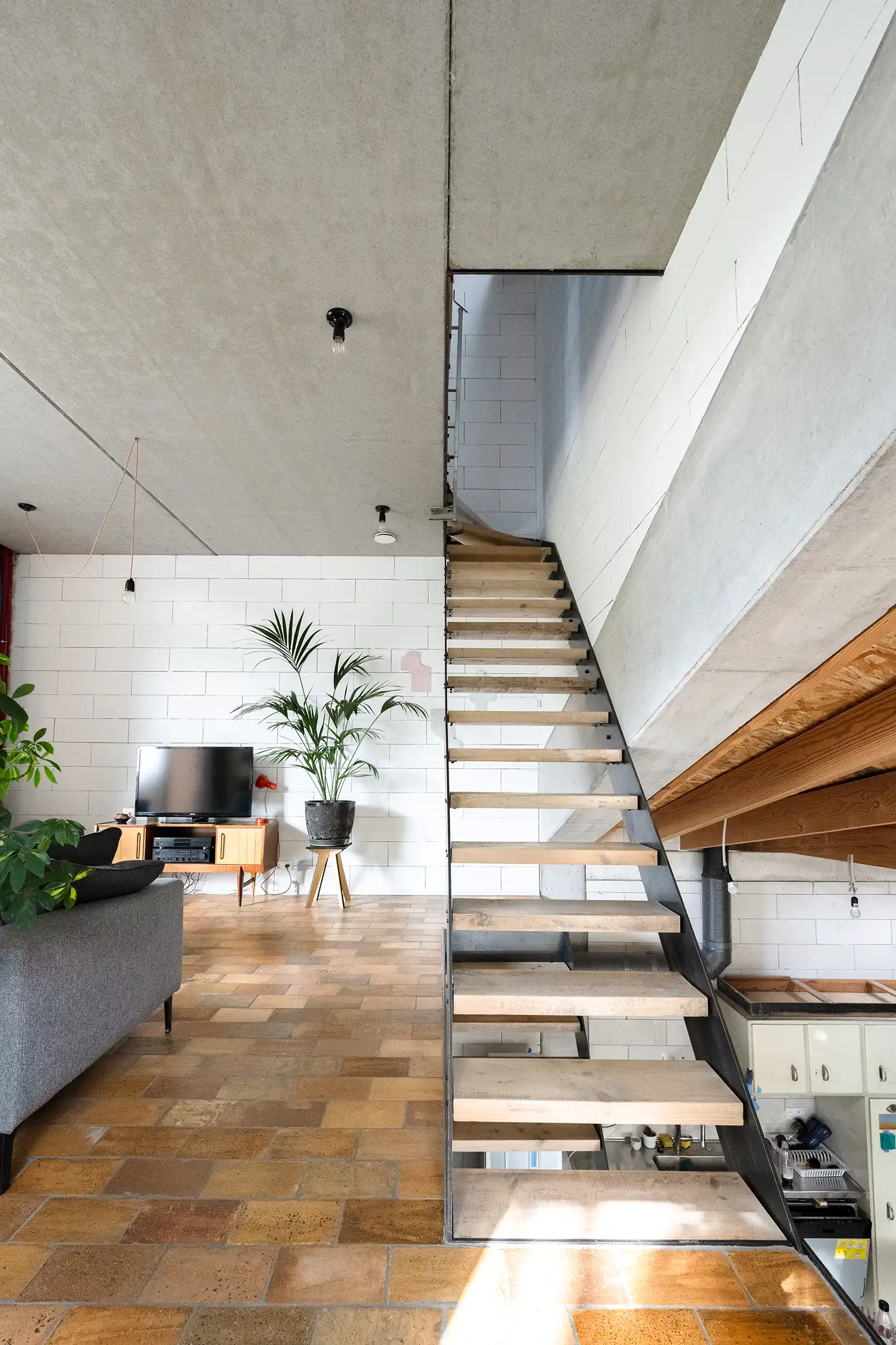 Interior view of a living space with exposed concrete ceiling and white brick walls, a dark metal and wood staircase leading upstairs, a grey sofa, and terracotta tiled floor.