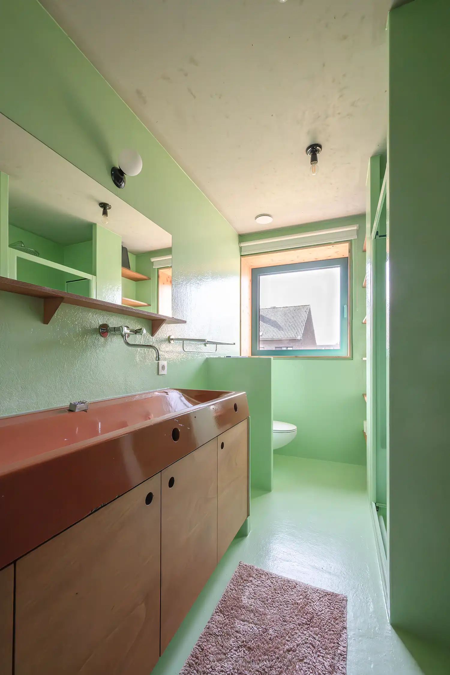 A bathroom with mint green walls and floor, a long wooden vanity with a reddish-brown sink, a square window with green frames, and a shower area.