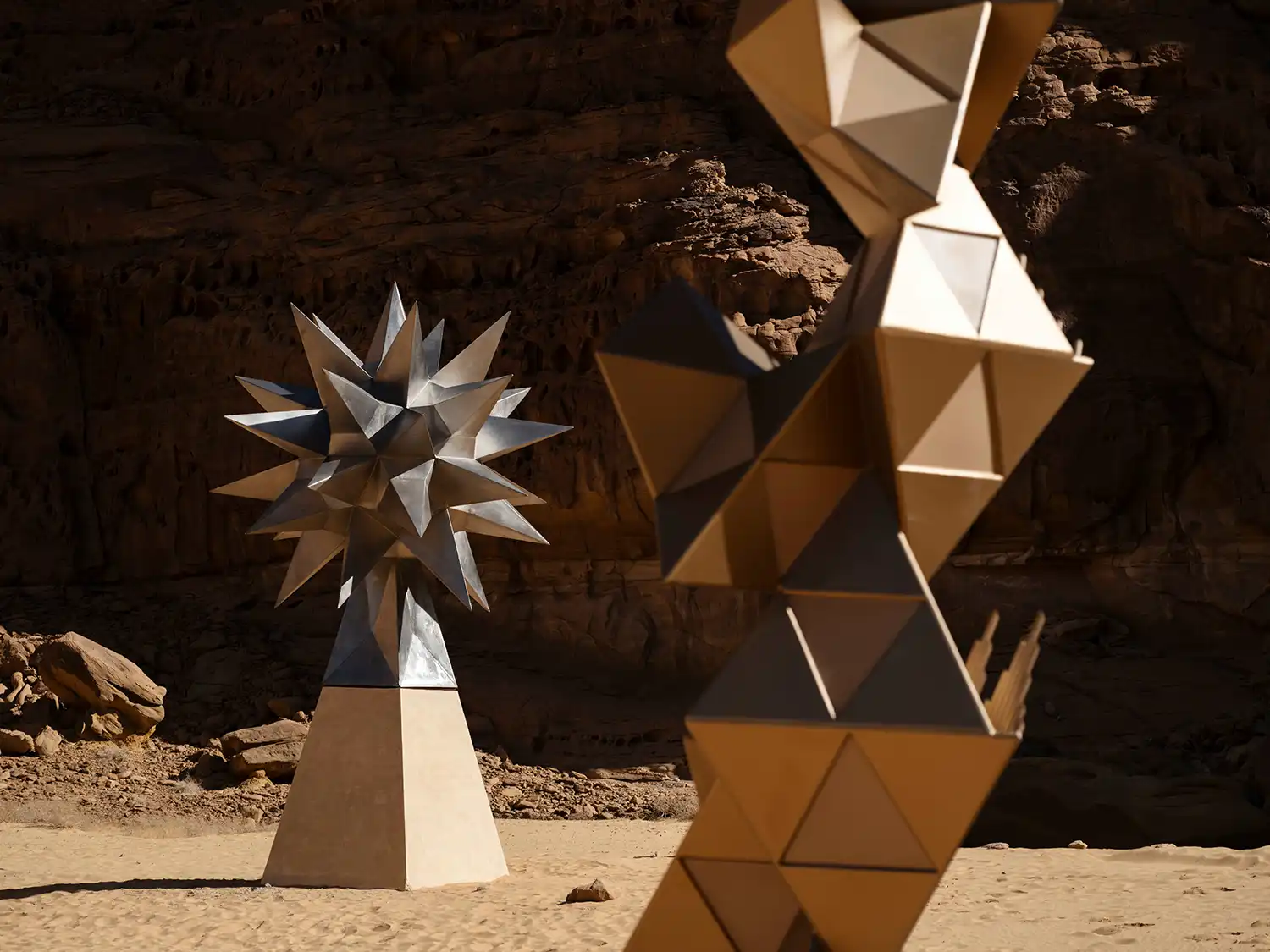 Two abstract geometric sculptures in metal and gold tones—one a multi-pointed star and the other a modular tower—standing in the AlUla desert.
