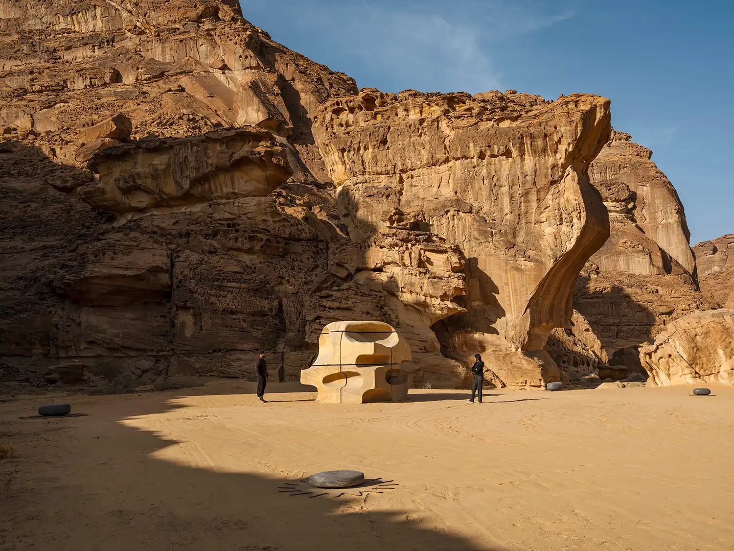 A large, tan-colored sculptural block with organic cavities by Basmah Felemban, backed by massive sandstone cliffs.