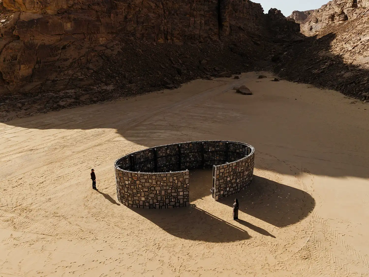 An oval-shaped stone enclosure within a metal mesh frame by Vibha Galhotra, casting a long shadow on the desert sand.
