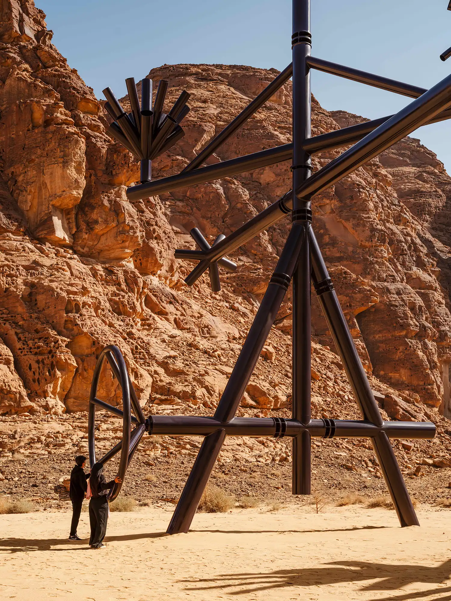 A tall, black kinetic sculpture by Bahraini-Danish with tubular arms, standing against a steep, sunlit sandstone cliff.