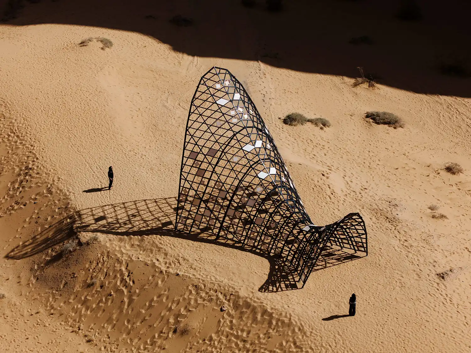 A large, triangular dark metal lattice structure by Héctor Zamora, casting a complex gridded shadow on a sand dune.
