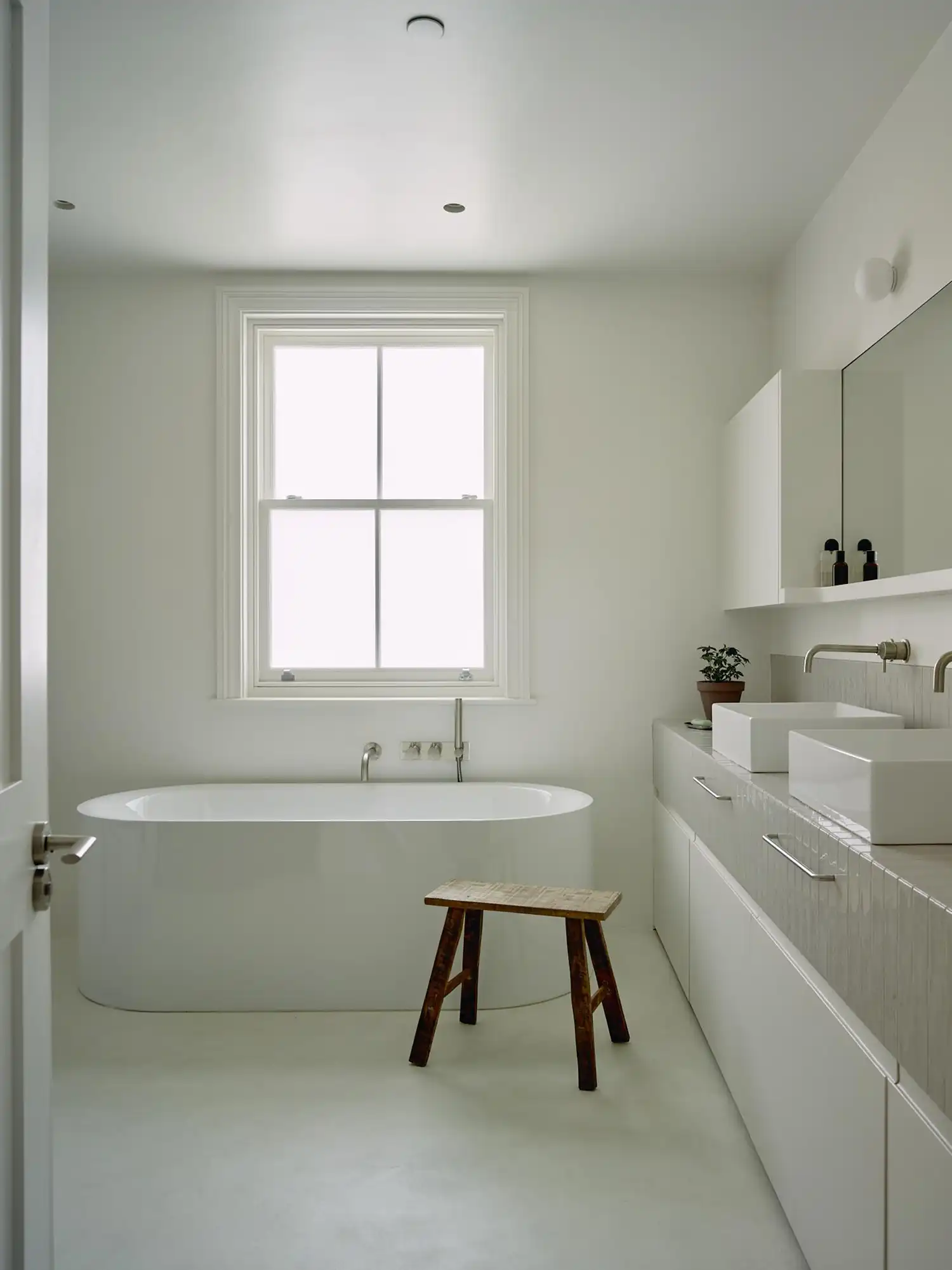 Minimalist white bathroom with a large soaking tub, double sinks, and a traditional sash window.