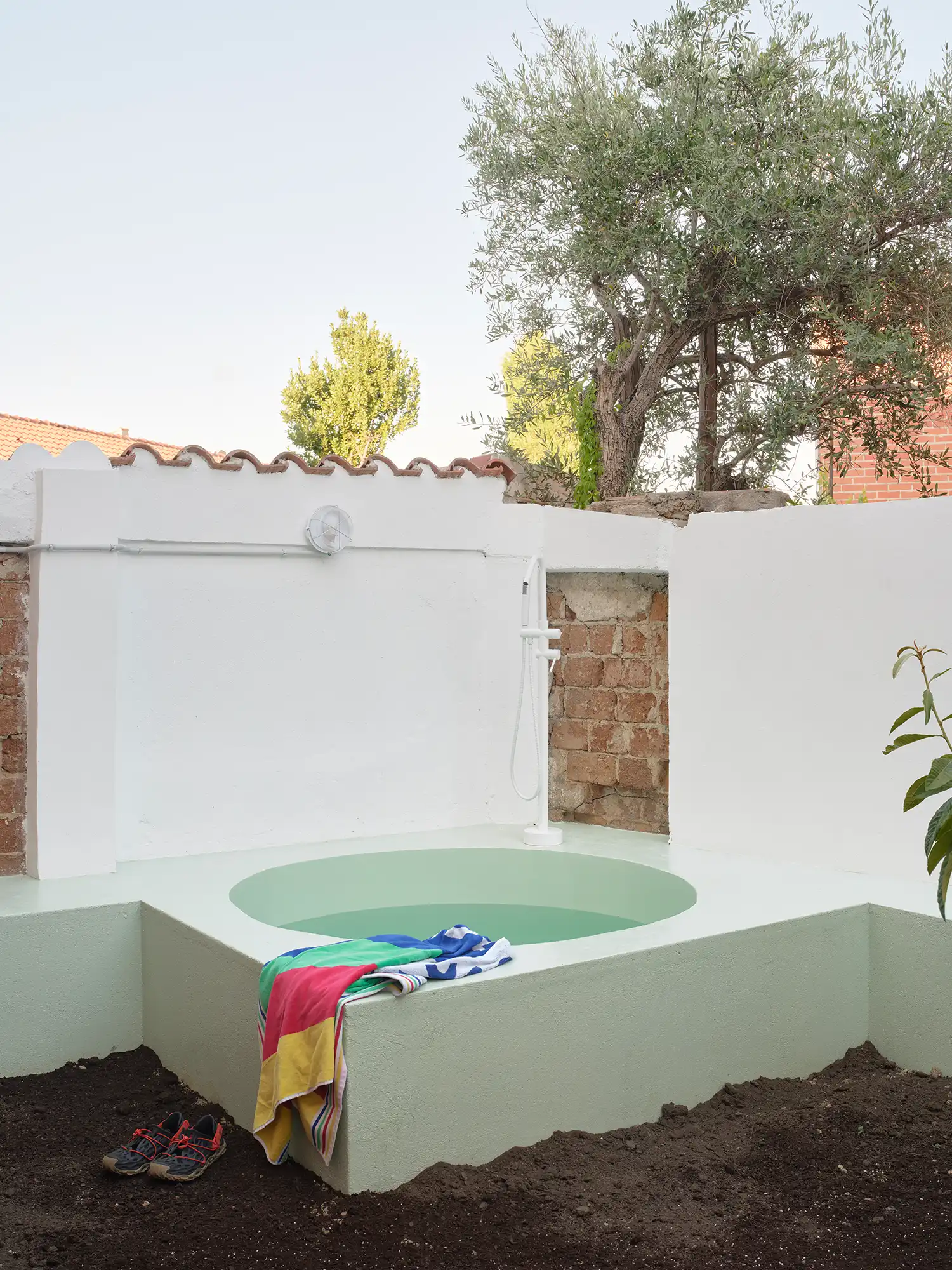 A sculptural mint green circular bathtub in a white-walled backyard with an olive tree and exposed brick.