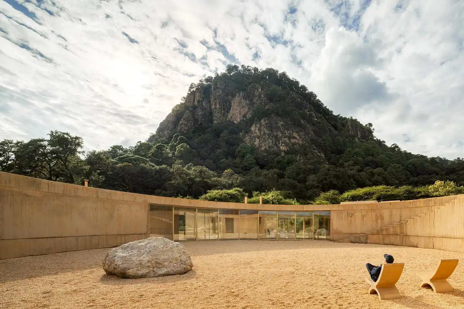 A person sitting in a wooden chair in a large circular gravel courtyard with a monumental rock and mountain view.