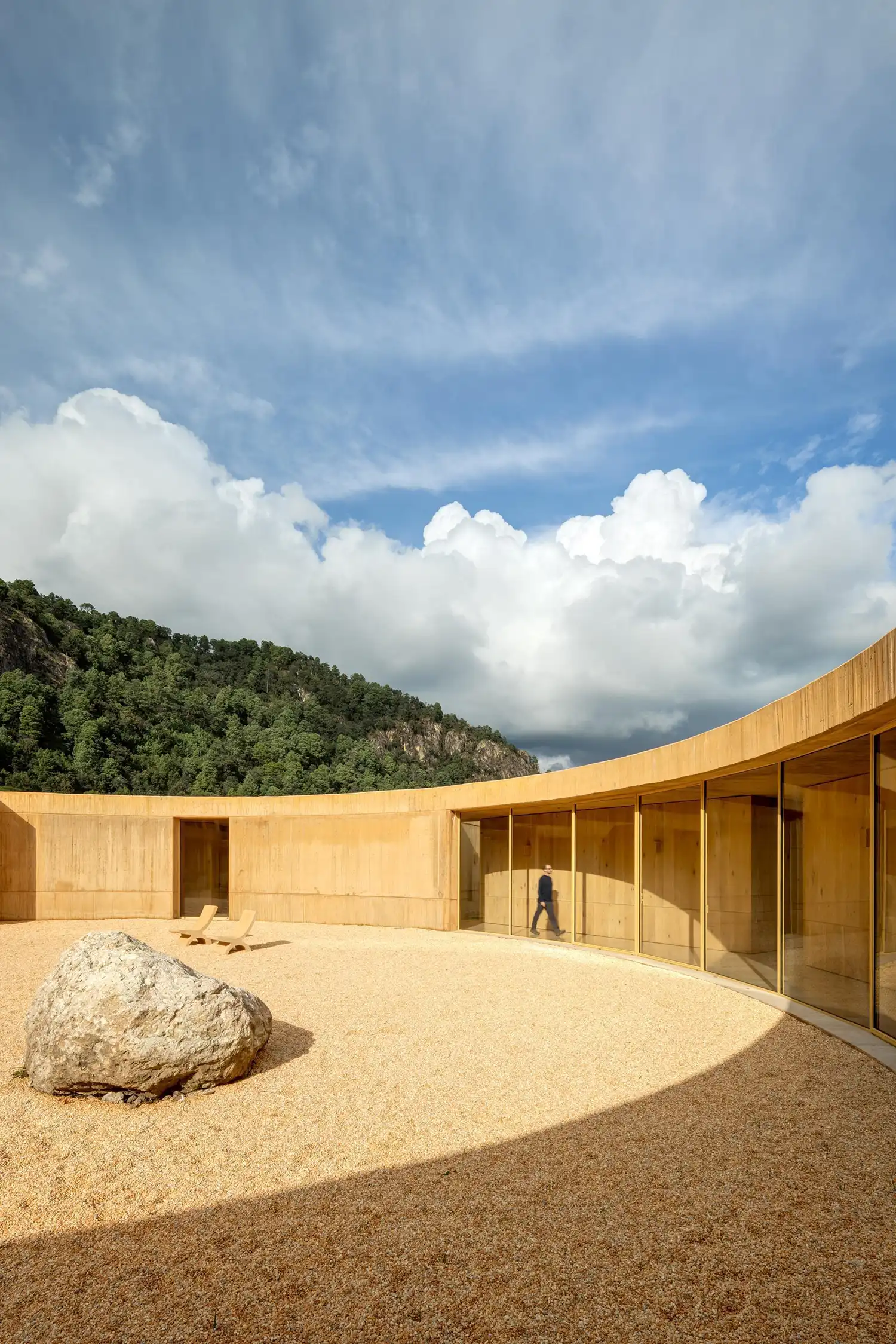 A person walking past a curved glass facade in a circular courtyard with a large rock on gravel.