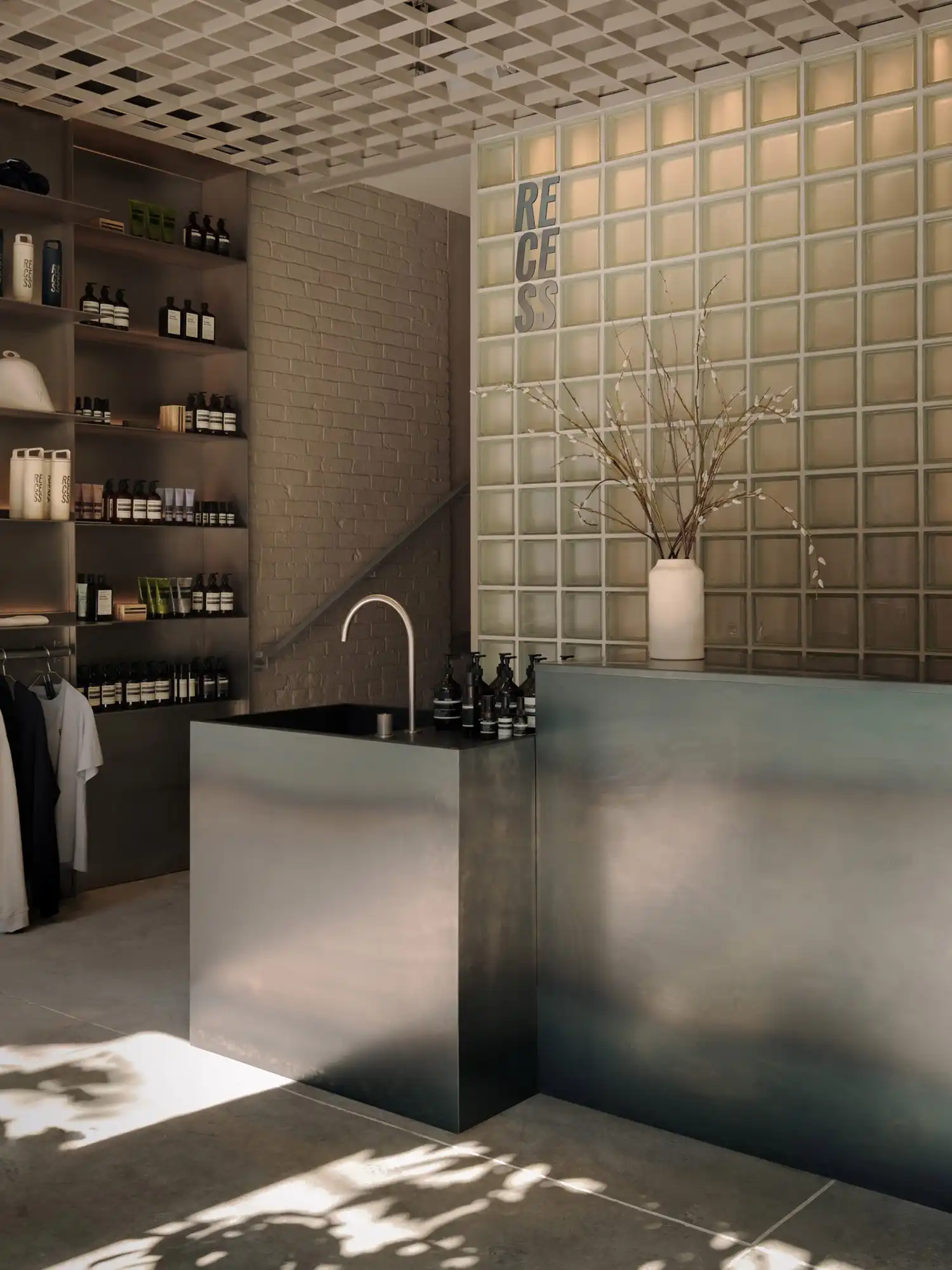 Industrial-chic reception area featuring a stainless steel desk, a glass block wall, and Aesop skincare products on metal shelving.