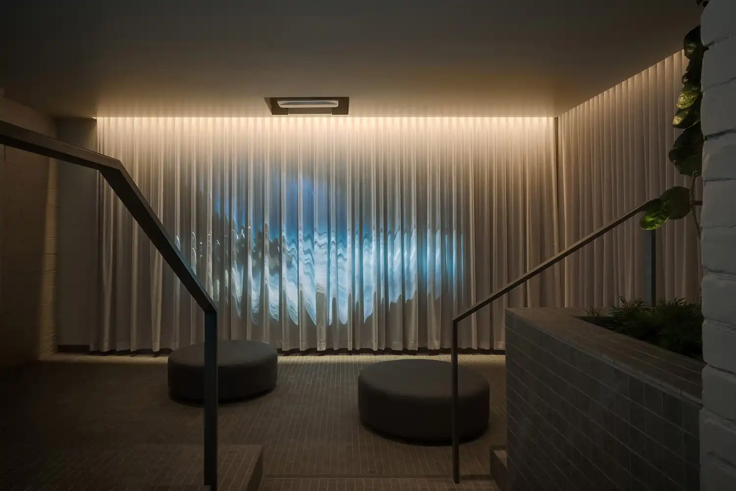A relaxation room with dark floor poufs and a large backlit curtain wall reflecting moving water ripples.