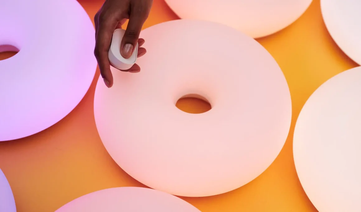 A close-up shot of a hand using a small white circular remote to control a matte pink donut-shaped lamp.
