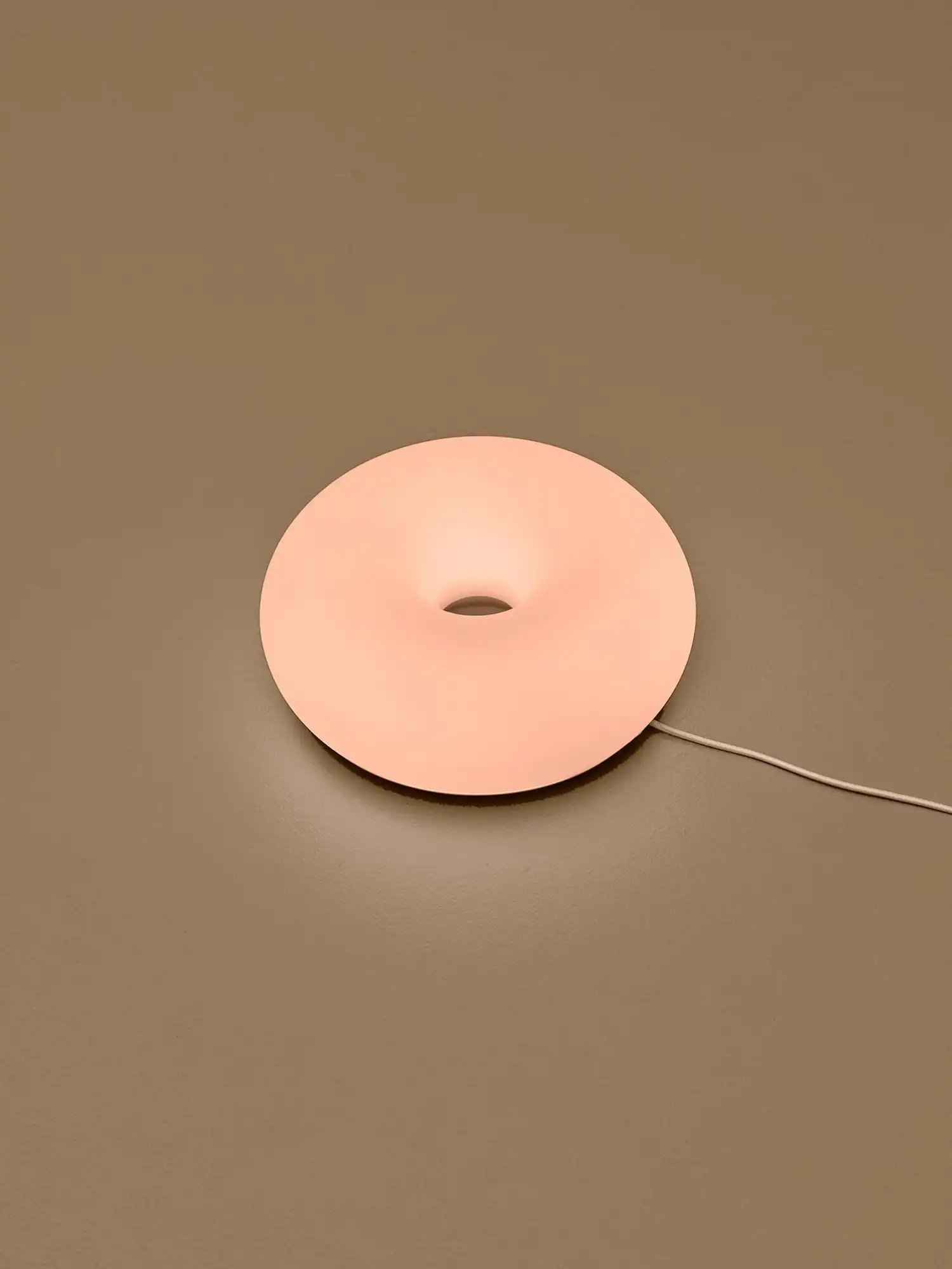 A single matte peach-colored donut lamp glowing on a flat brown surface with its power cord extending to the right.