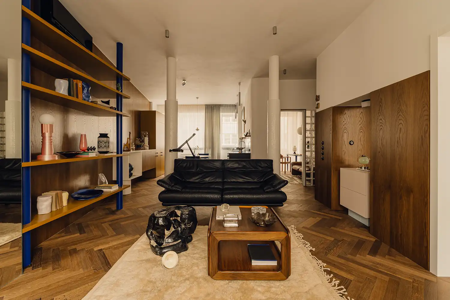 Open-plan living area with a black leather sofa, blue shelving, and white tiled columns.