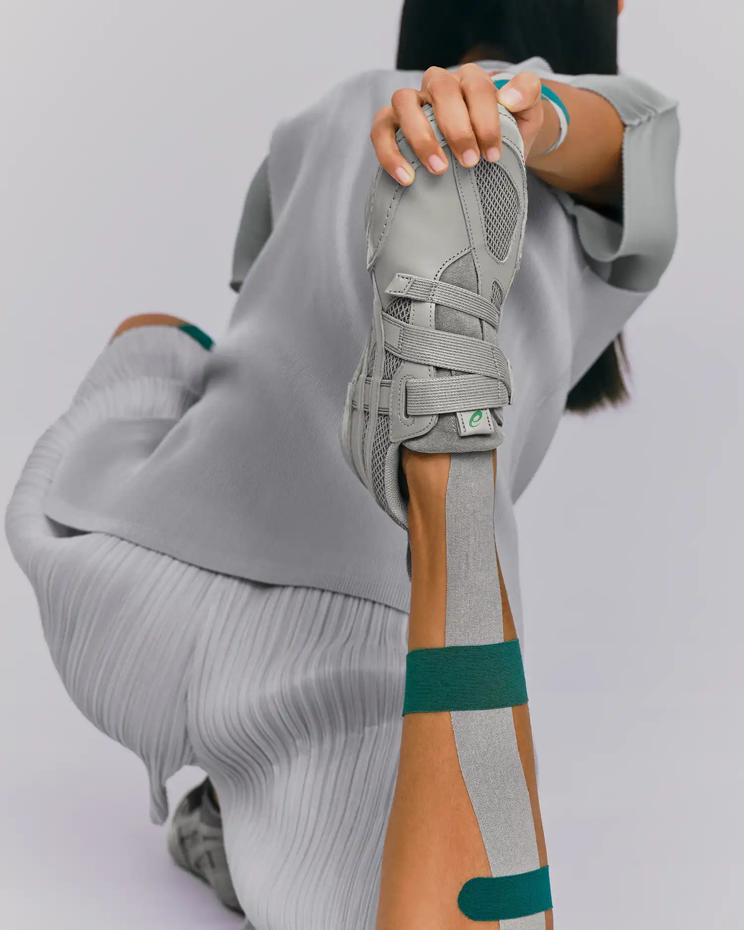 A model in grey pleated apparel holding a matching HYPER TAPING™ sneaker during a stretching pose to demonstrate flexibility.