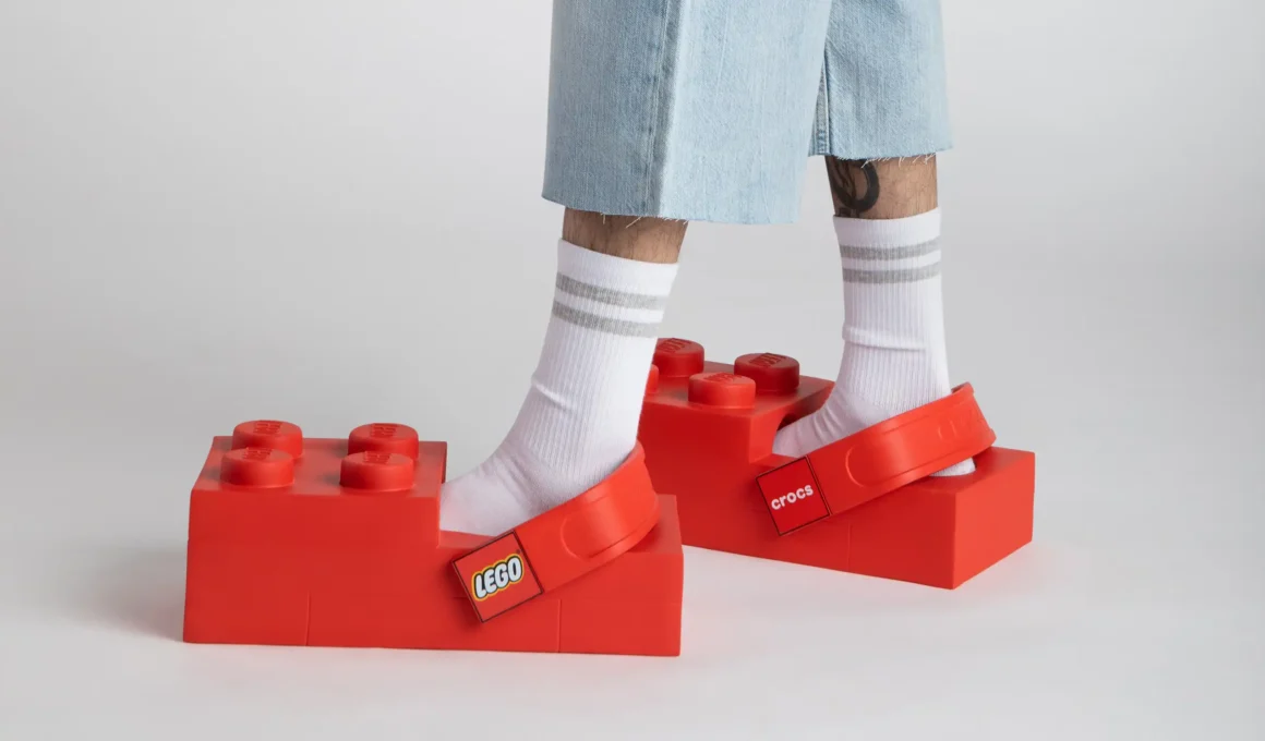 A person wearing oversized red LEGO Brick Clogs with white striped socks and denim shorts.