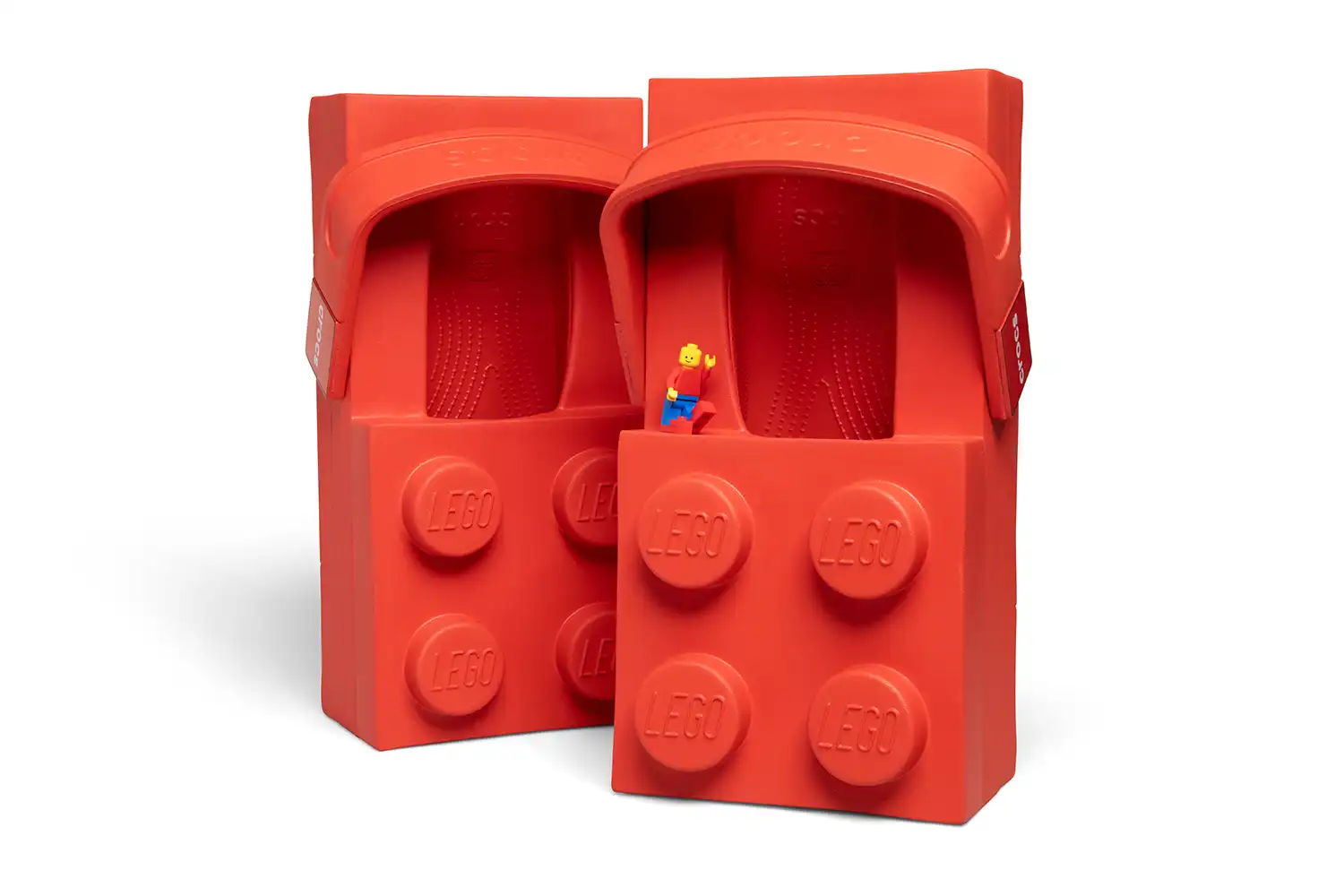 Two red LEGO Brick Clogs standing vertically next to a tiny LEGO Minifigure for scale.