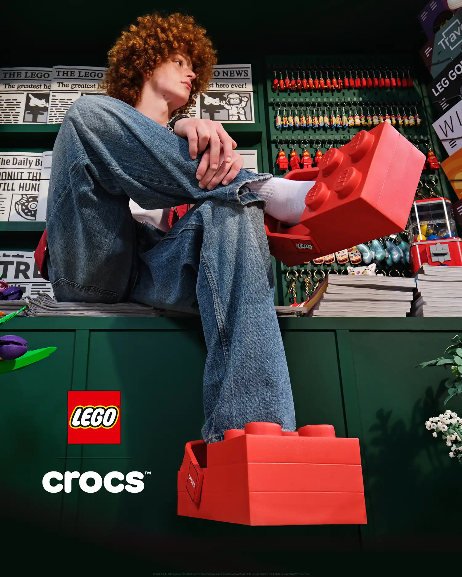 A person with curly hair sitting in a LEGO-themed environment wearing the red LEGO Brick Clogs.