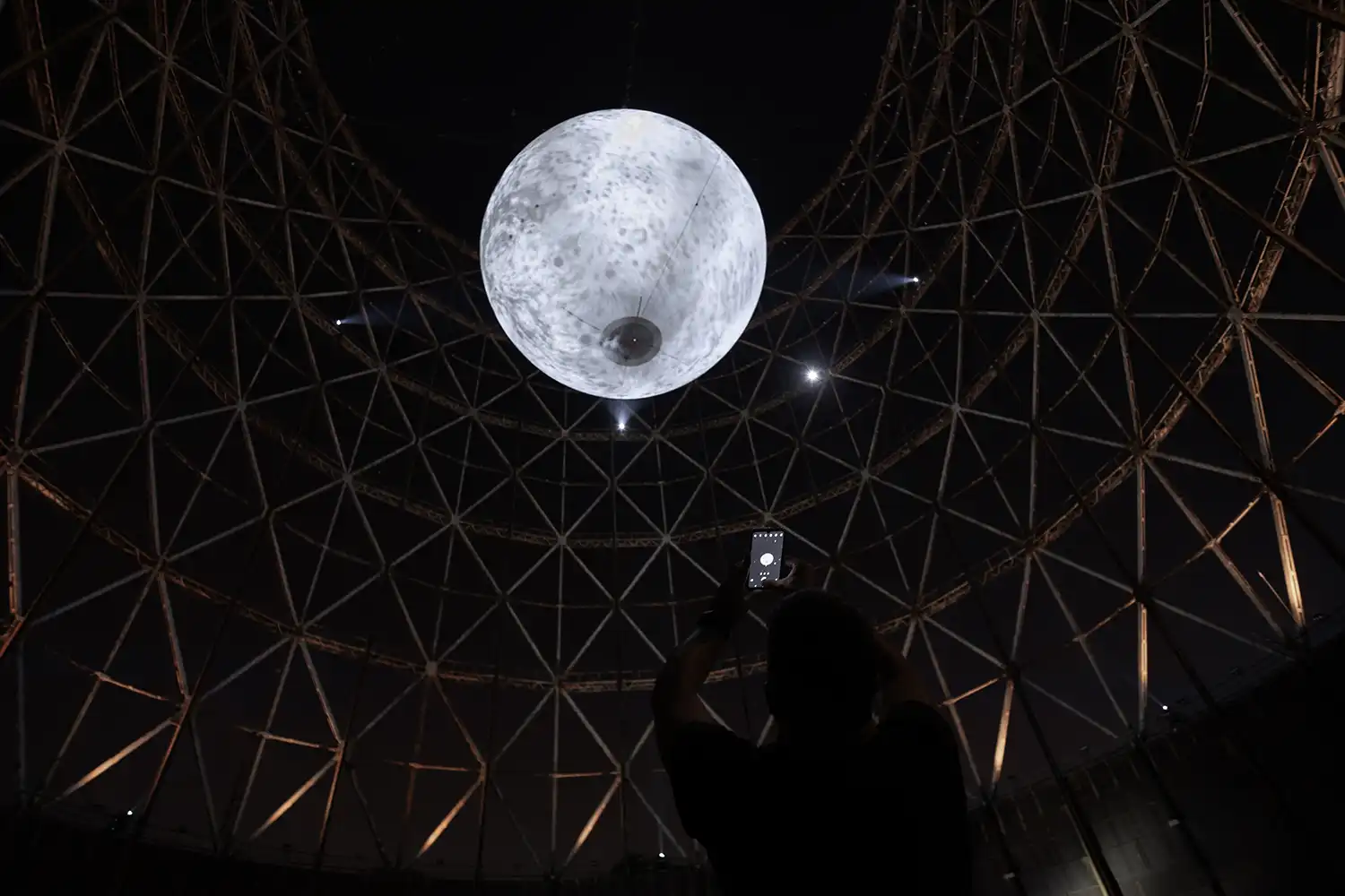 A person in silhouette holding up a smartphone to photograph the glowing lunar installation inside the dark lattice structure.