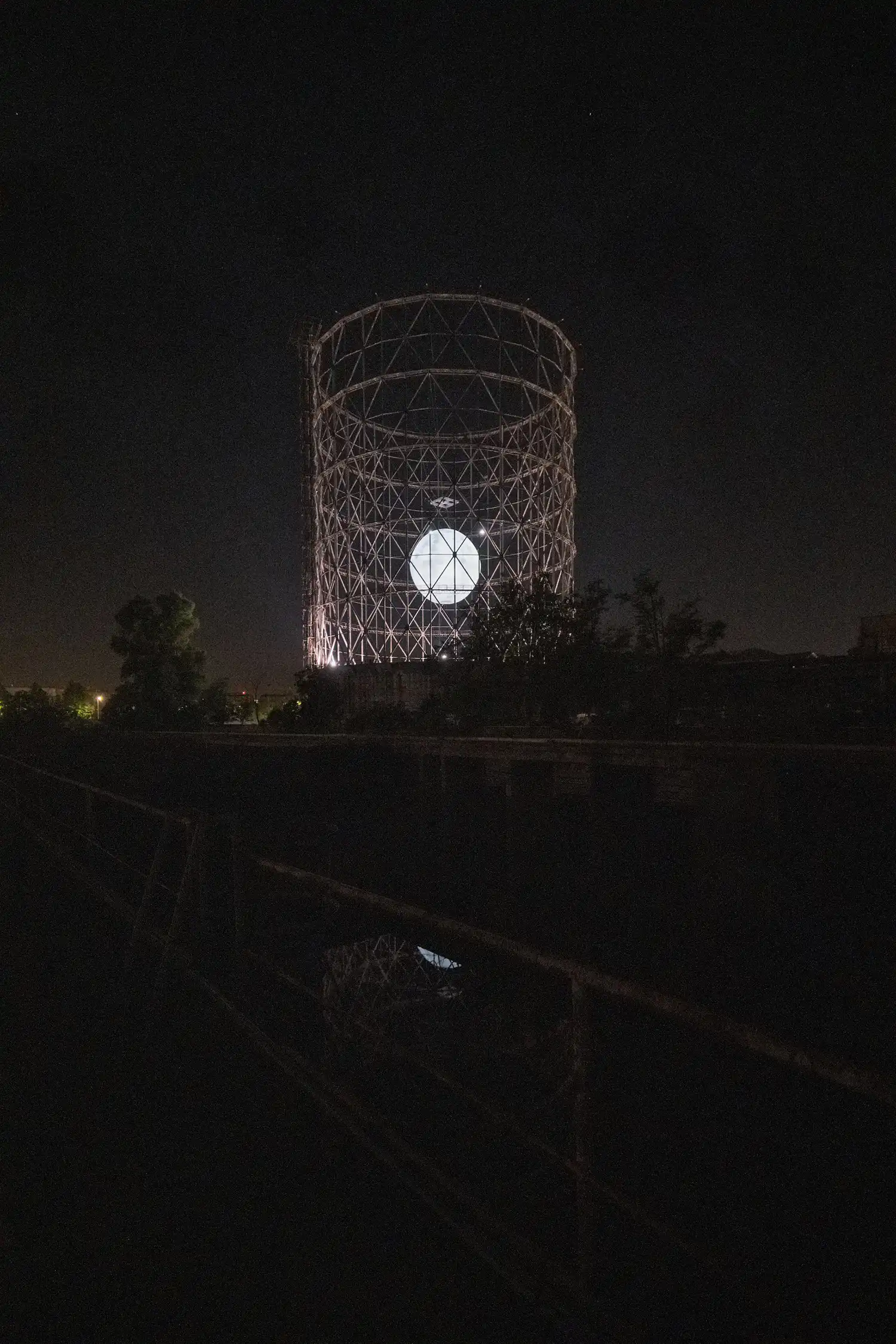 A distant night view of the illuminated Gasometer reflecting in the dark water of a foreground canal.