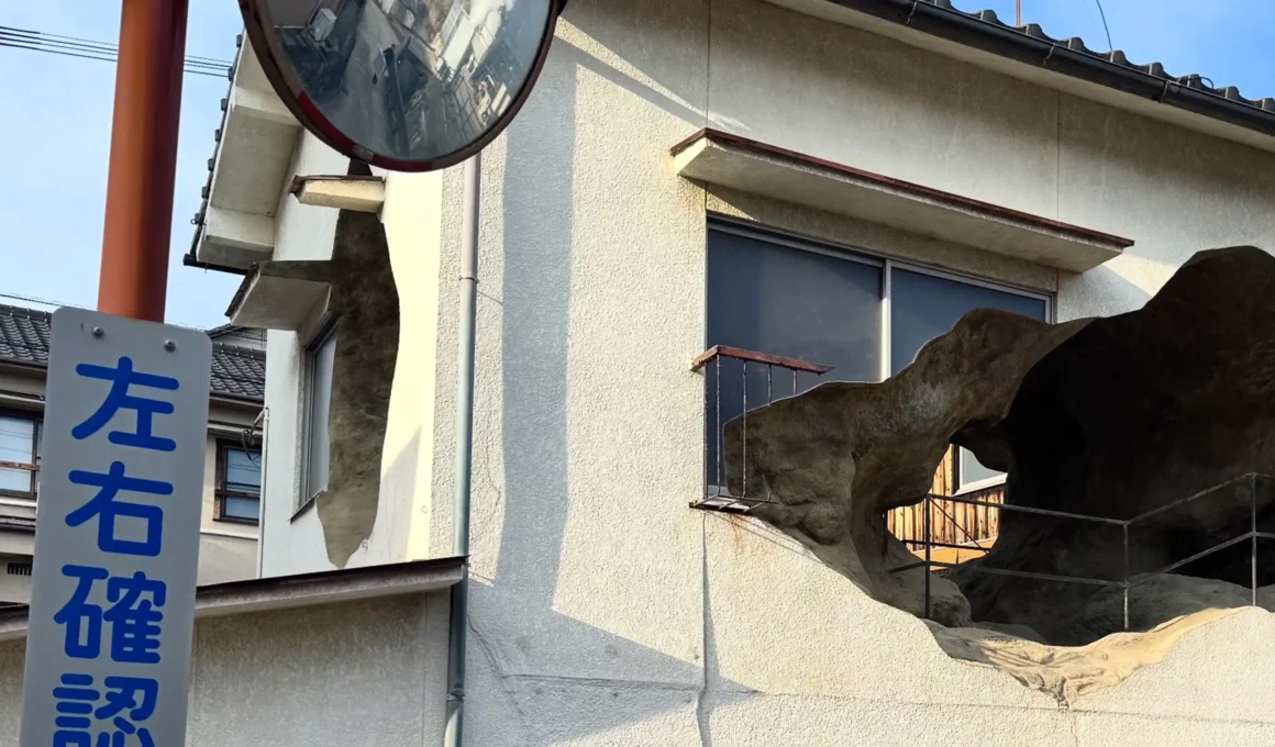 Exterior view of a white two-story Japanese house with large, irregular jagged holes cut into the facade revealing a rock-like interior.
