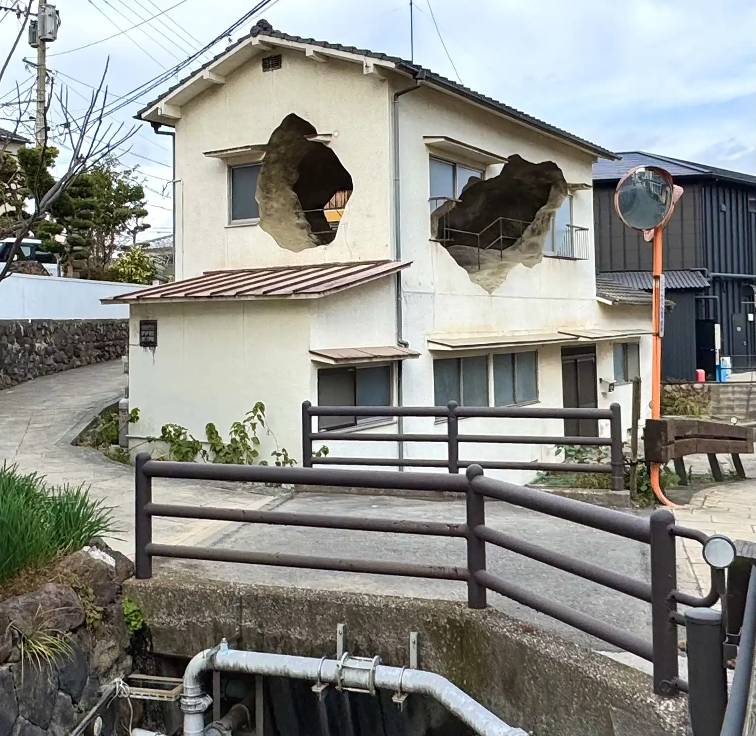 Wide shot of a traditional house in a Japanese neighborhood with large circular excavations in its walls and a black metal staircase leading to one.