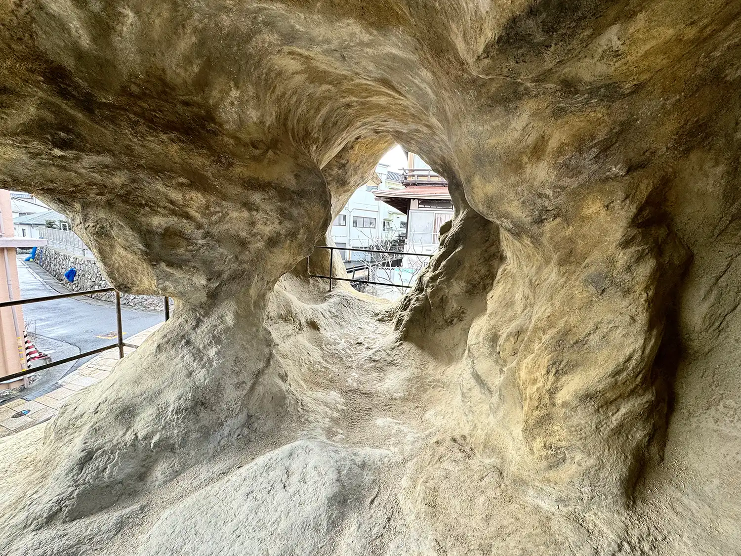 Interior view of a cave-like space with rough, beige textured walls and an irregular opening looking out onto a Japanese street.