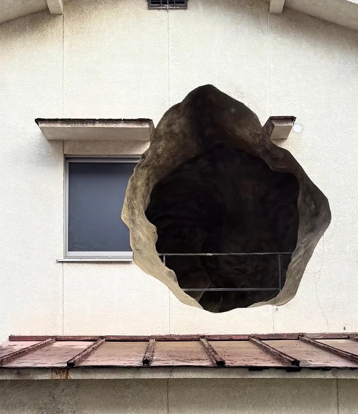 Close-up of a large, dark, irregular hole in a white building wall next to a standard rectangular window.