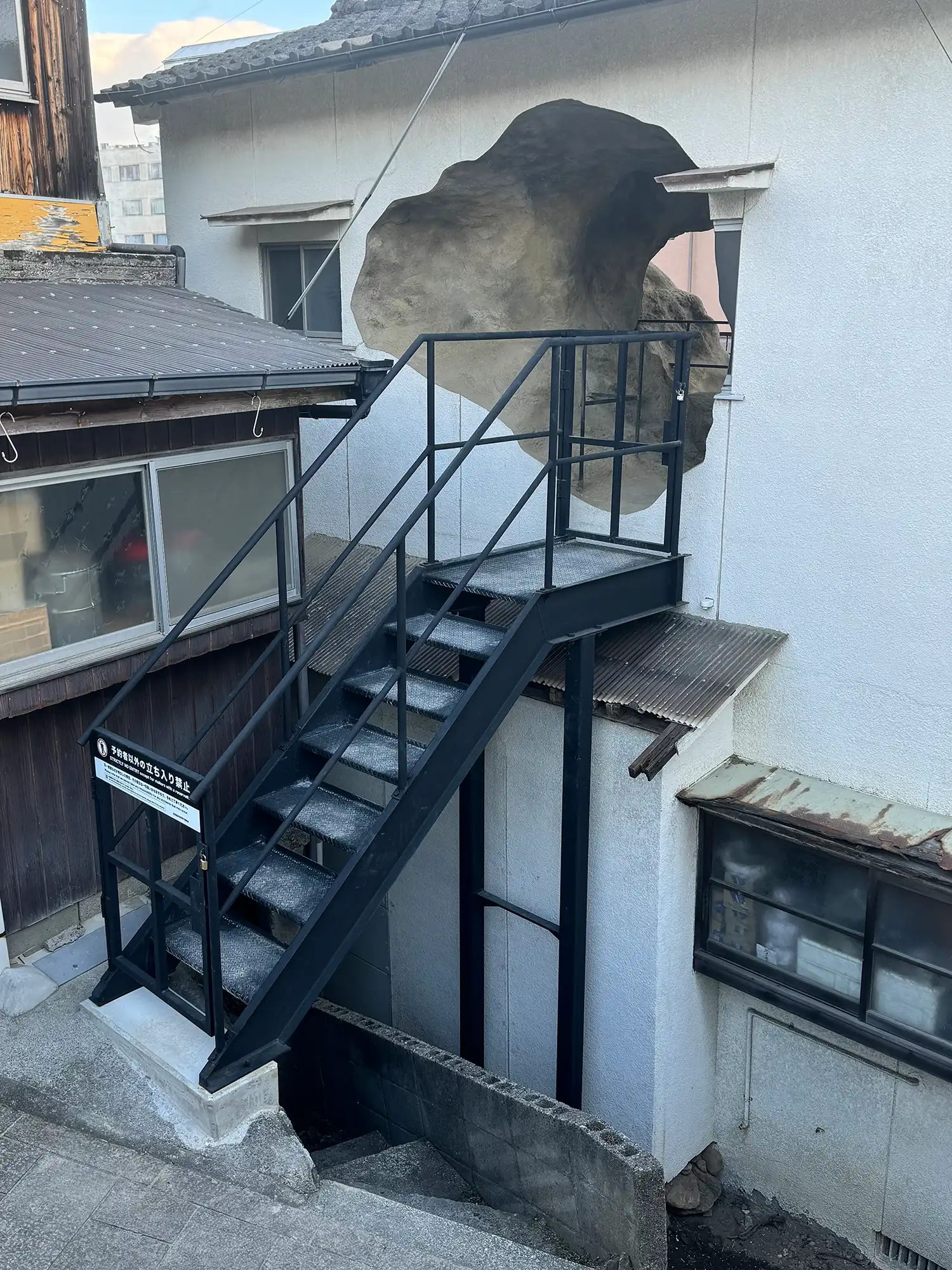 A black industrial metal staircase leading up to a large jagged hole in the side of a white house.