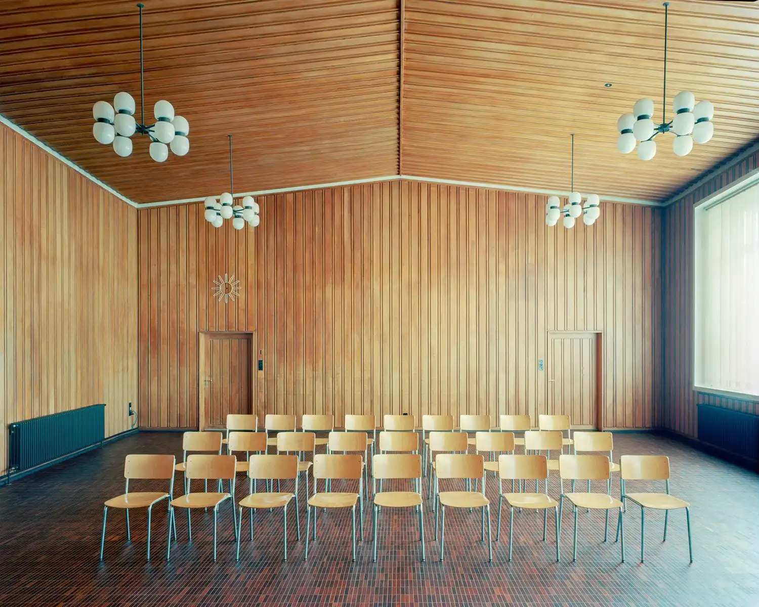 Mid-century assembly hall with vertical wood cladding and bubble chandeliers.