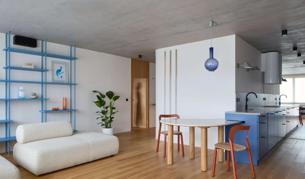Wide view of an open-plan apartment in Prague featuring raw concrete ceilings, light oak wood floors, and a blue kitchen island with a curved marble dining table.