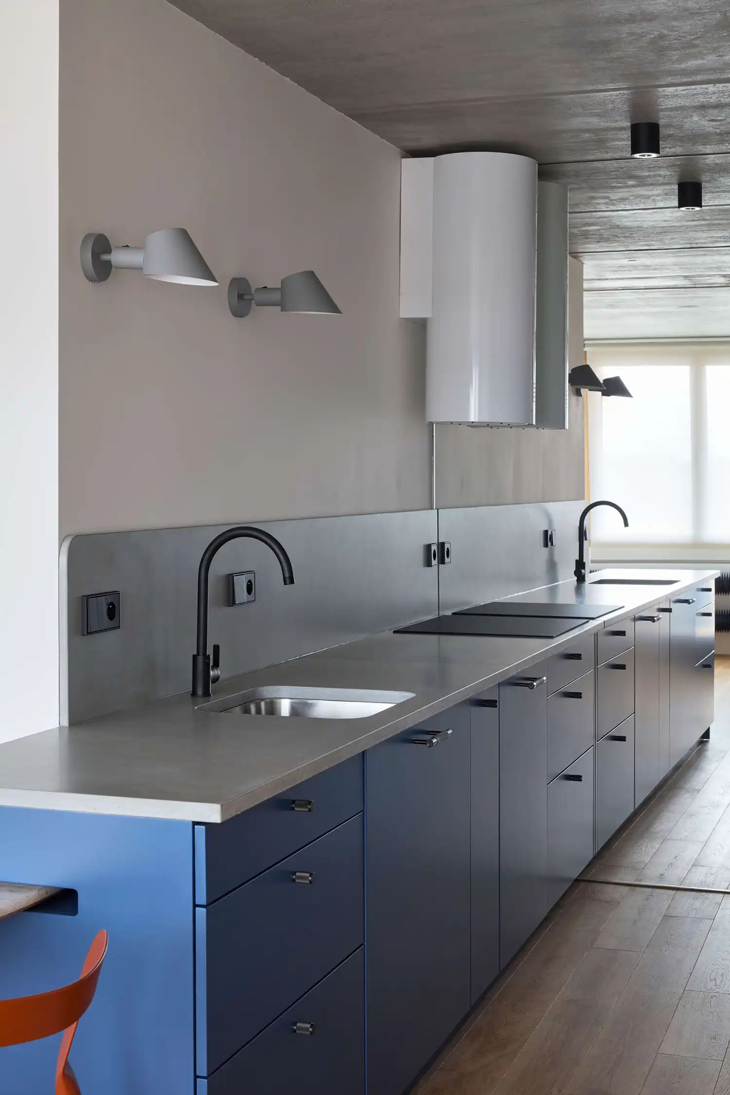 A long, minimalist kitchen galley with blue cabinetry, a grey countertop, and black faucets beneath a concrete ceiling.