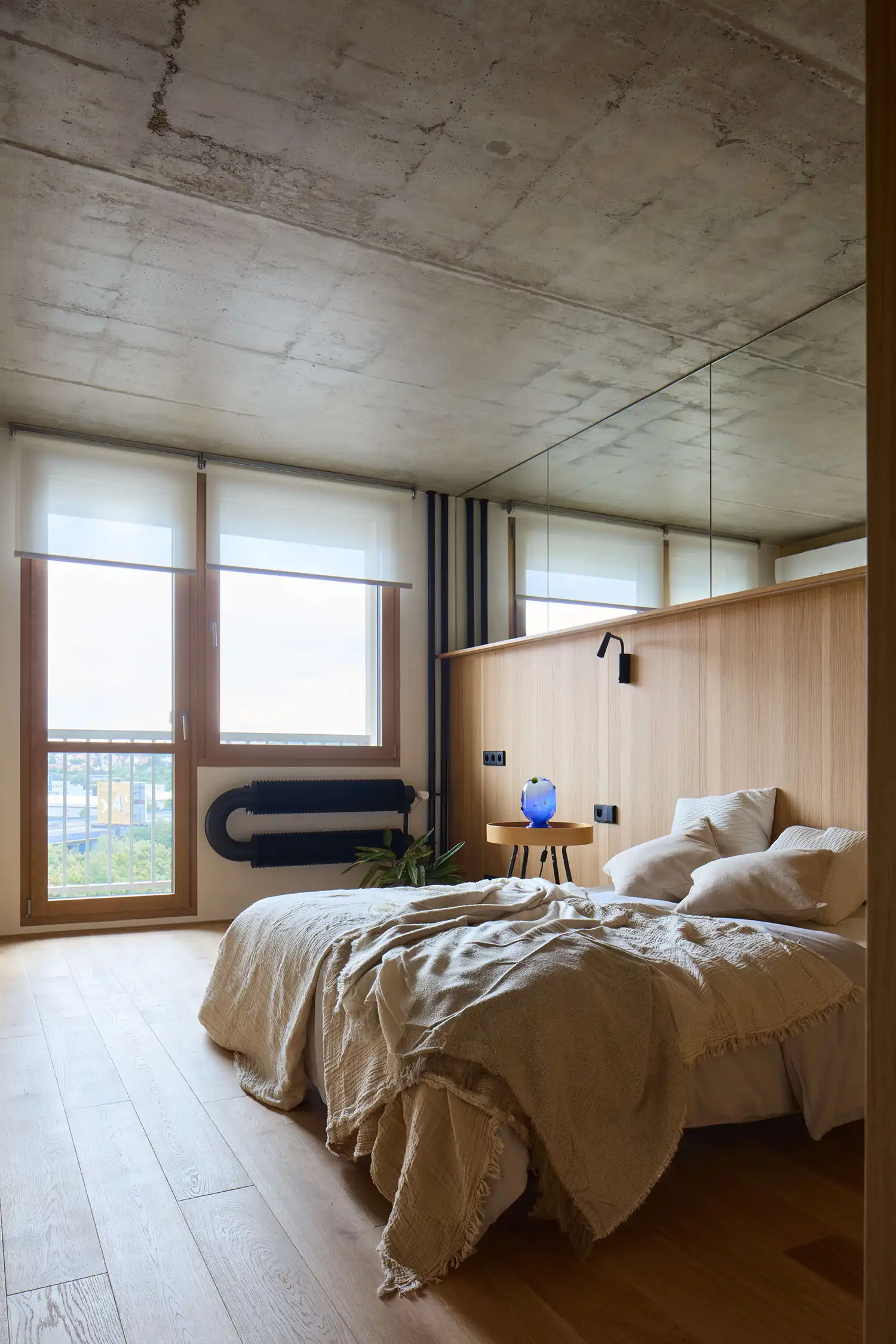 Minimalist bedroom in a Prague apartment with a large oak headboard, mirrored wall, and views of the city skyline.