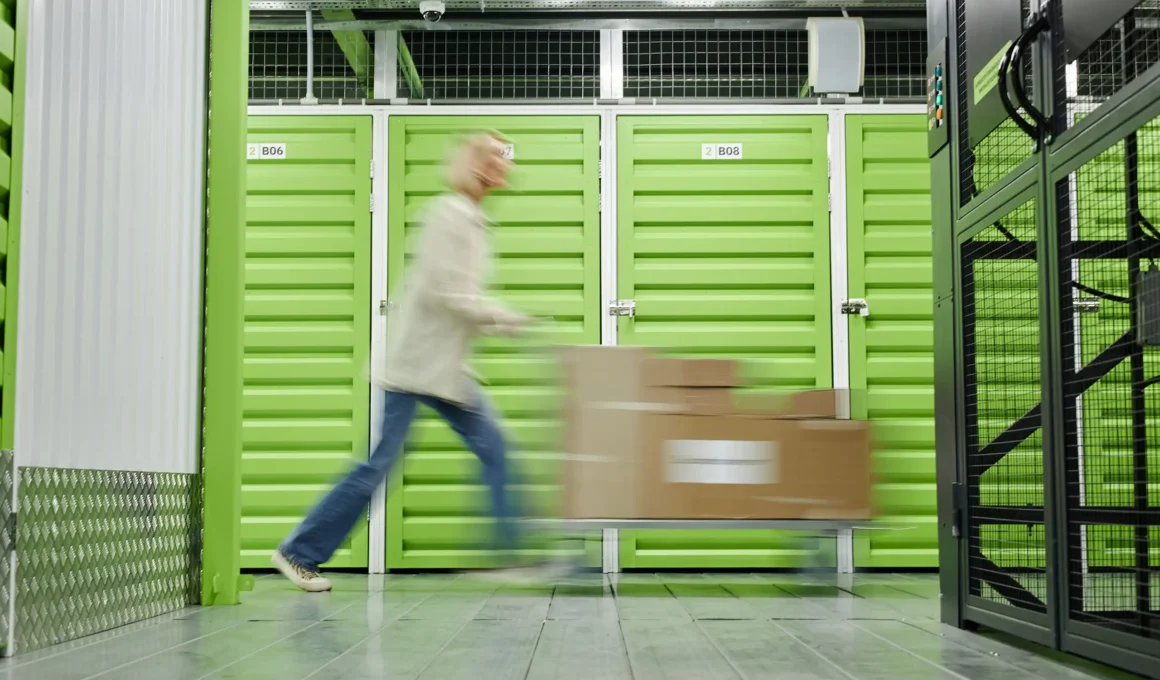 A person in motion blur pushing a trolley with boxes past bright green self-storage units in a modern industrial facility.