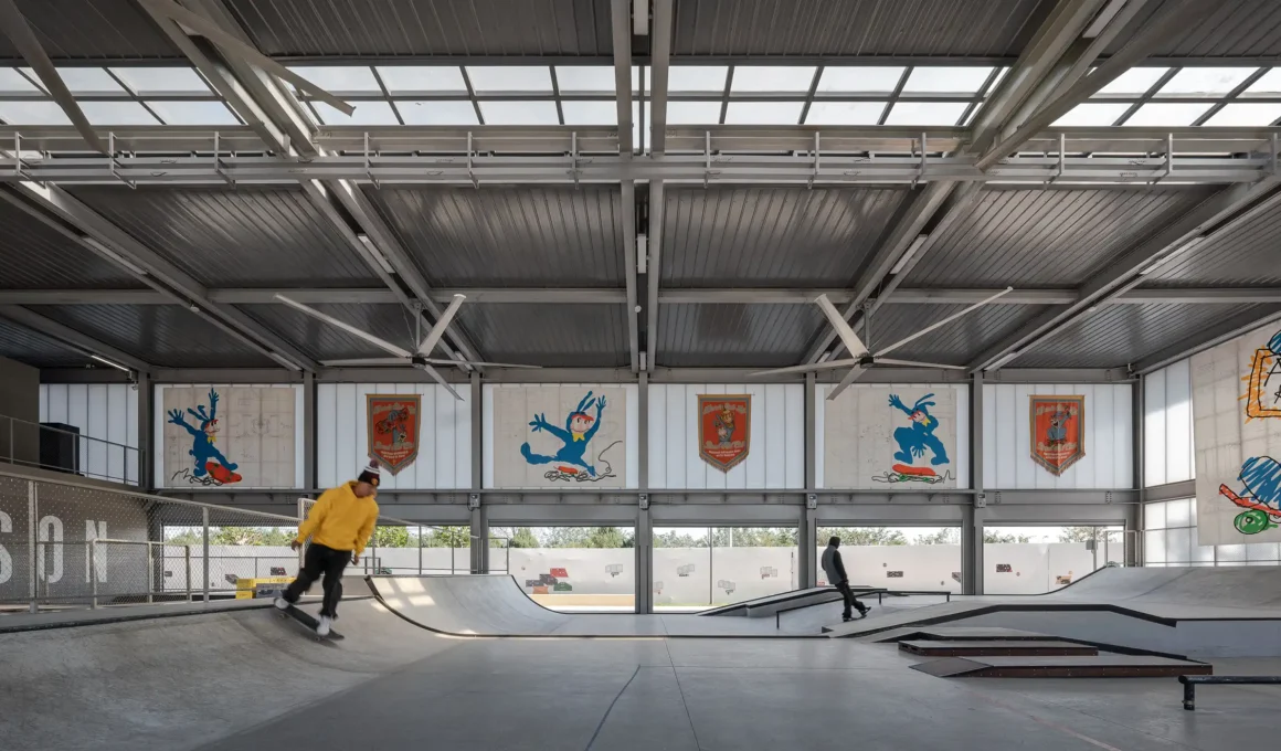 Professional skateboarders utilizing the indoor skatepark at AVENUE & SON North Coast, featuring high-span steel architecture, translucent polycarbonate facades, and industrial ceiling fans in Riverain, Aranya.