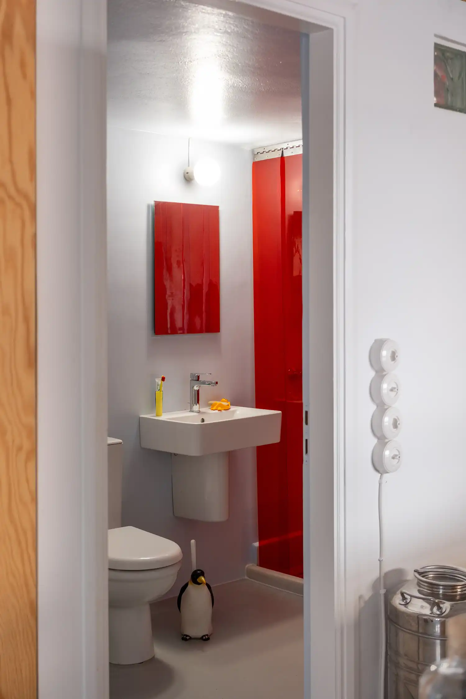 Minimalist bathroom interior viewed through a doorway, featuring a vibrant red shower curtain and retro switches.