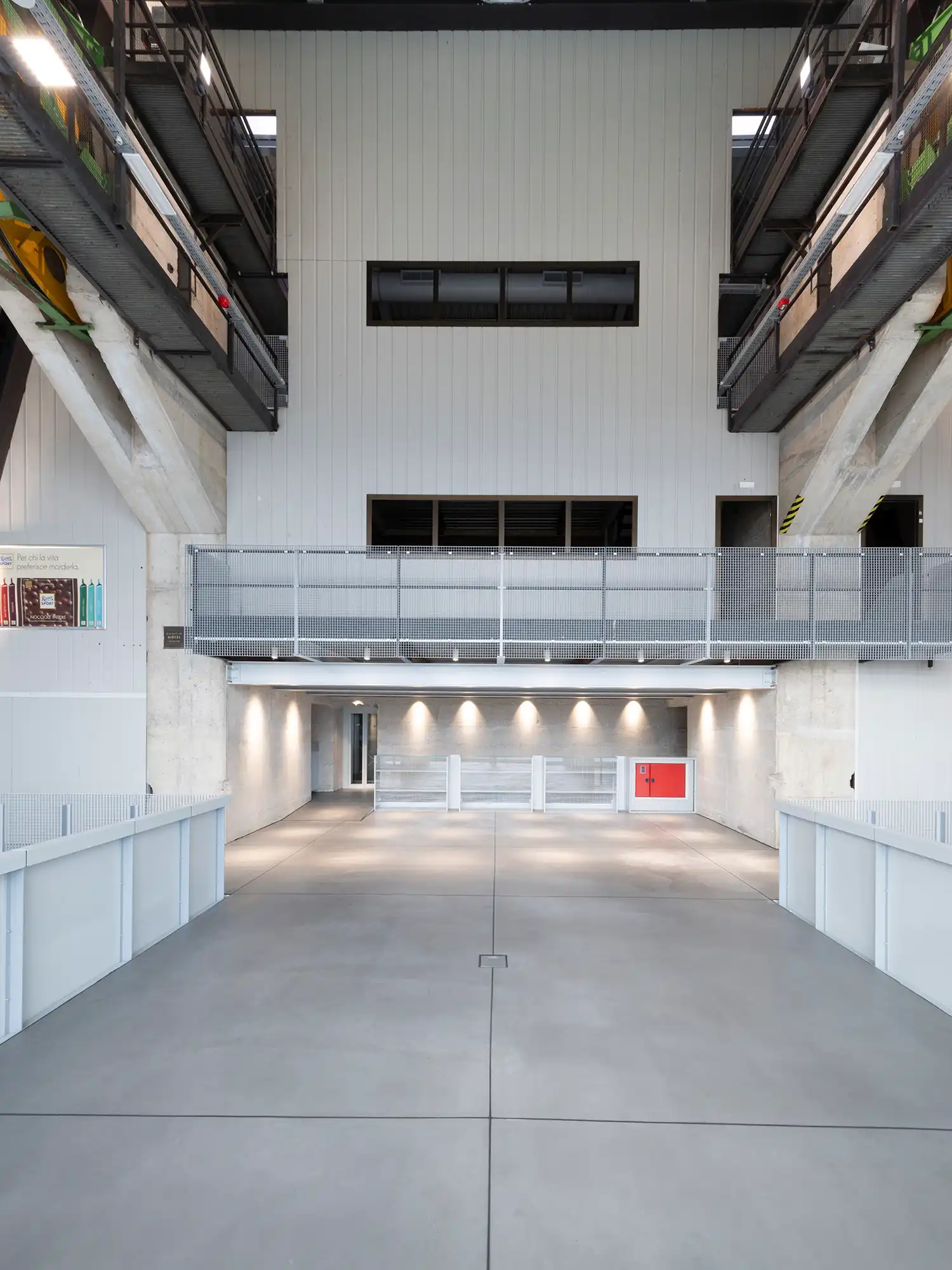 High-ceilinged industrial interior with white paneled walls and a metal mesh mezzanine bridge.