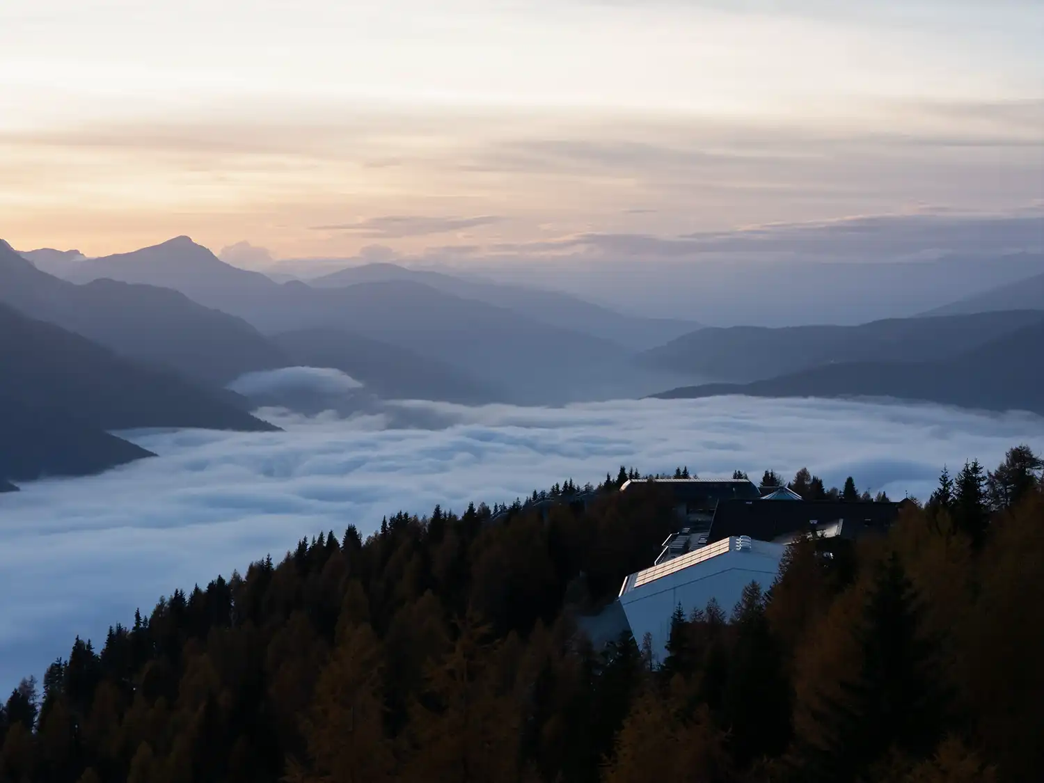 Aerial view of the Reinhold Messner Haus roof at sunset, overlooking a sea of clouds in the Italian Alps.