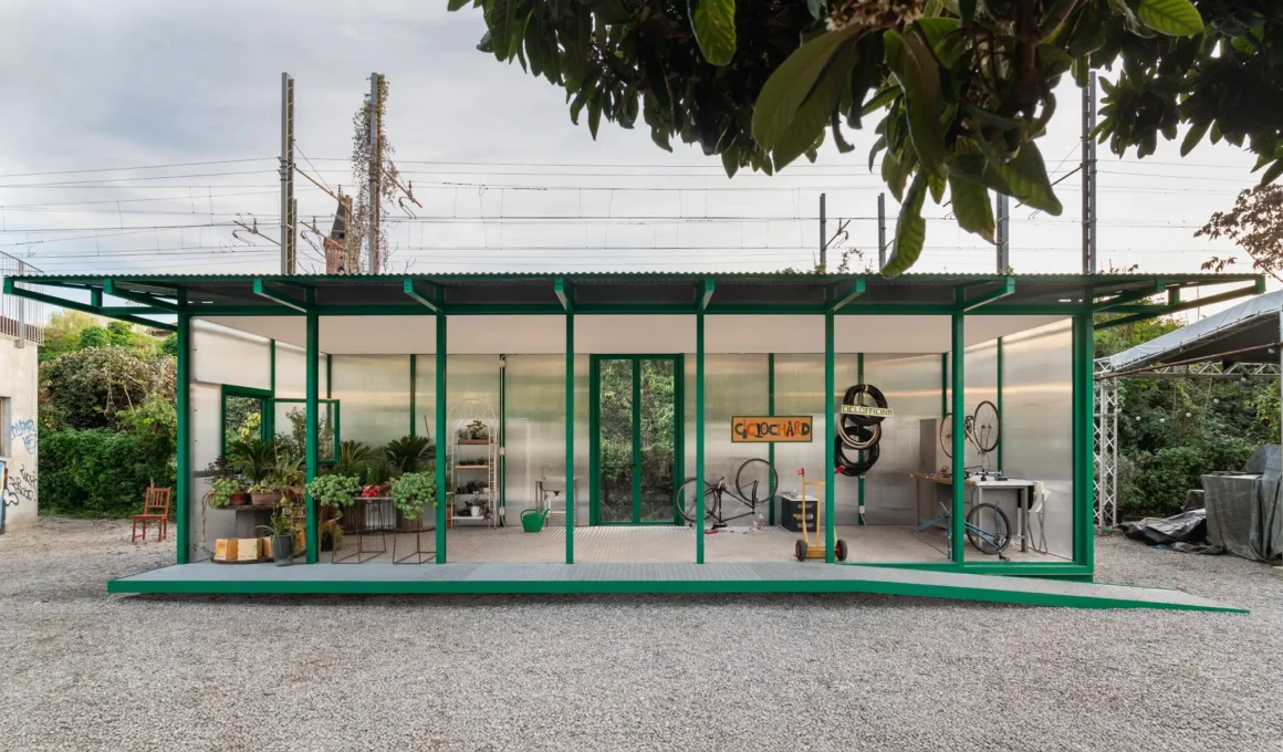 Wide shot of the San Cristoforo Ciclofficina, a green steel and polycarbonate pavilion in Milan.