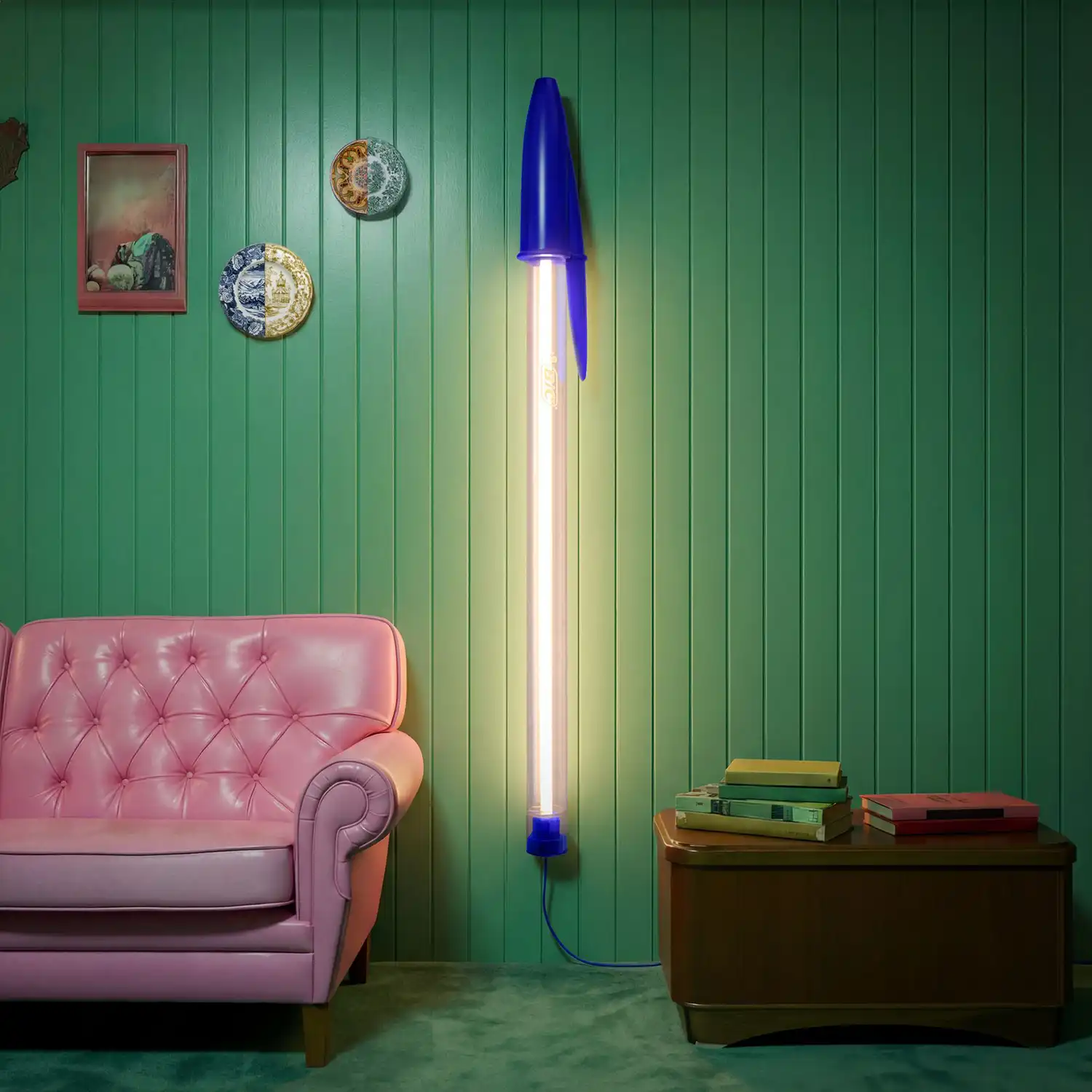 A blue Bic Cristal wall lamp mounted vertically on a green wood-paneled wall next to a pink sofa.