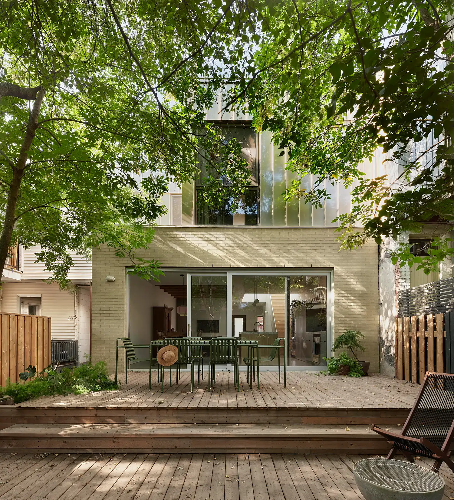 The rear facade of ShoeBox CHB in Montreal featuring large sliding glass doors, a wooden terrace, and a stainless steel upper volume nestled among trees.