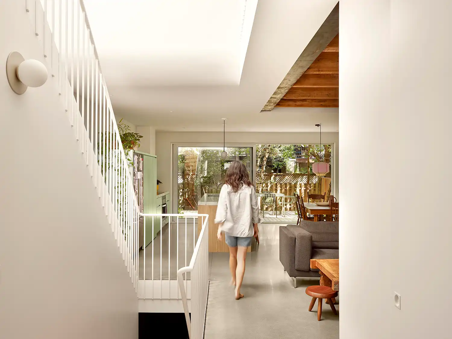 A view from the hallway into a light-filled open-plan living area featuring a central white staircase, wooden accents, and a garden view.