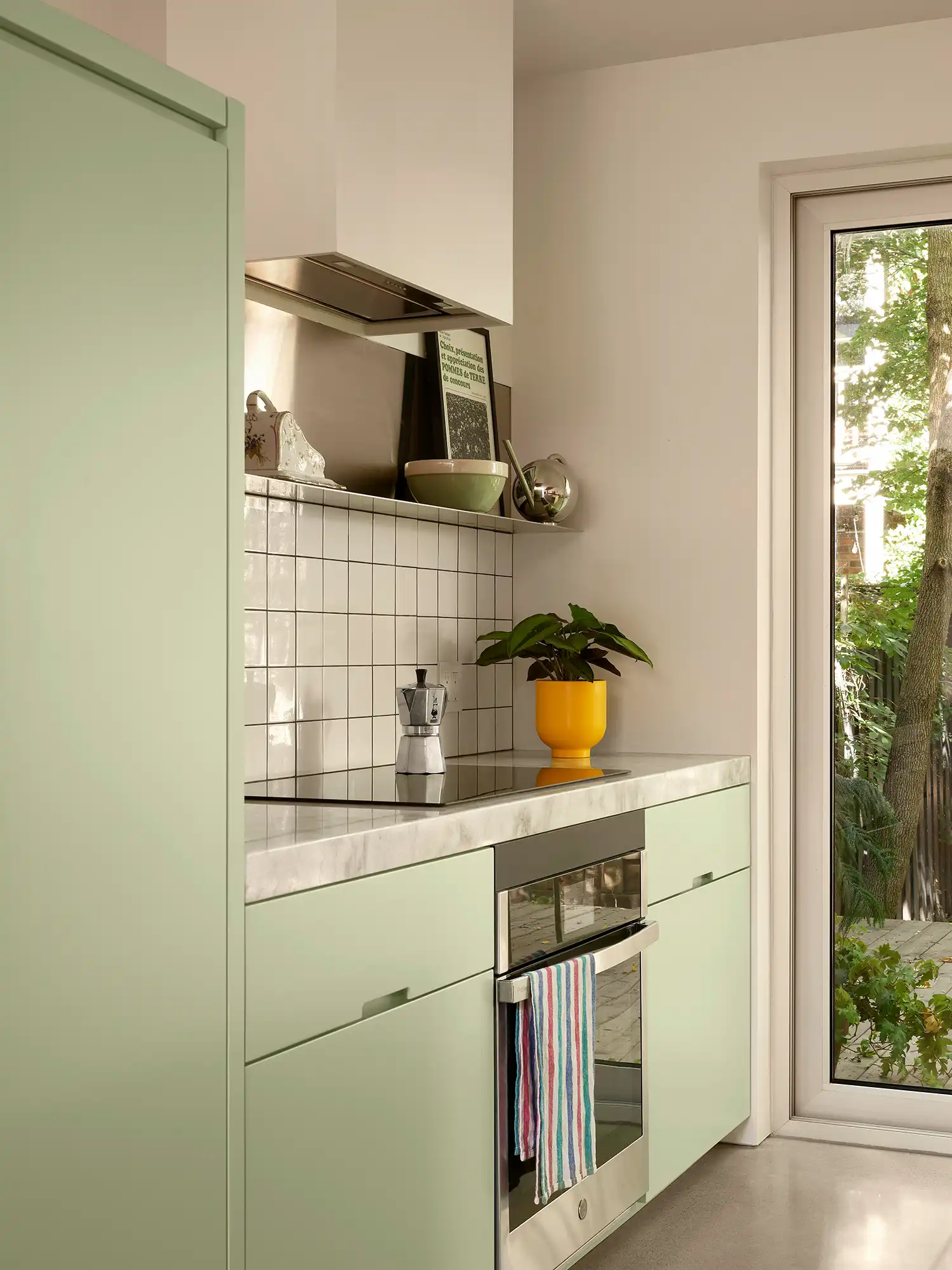 A modern kitchen detail in ShoeBox CHB featuring mint green cabinetry, marble countertops, white square tiles, and a view of the backyard.