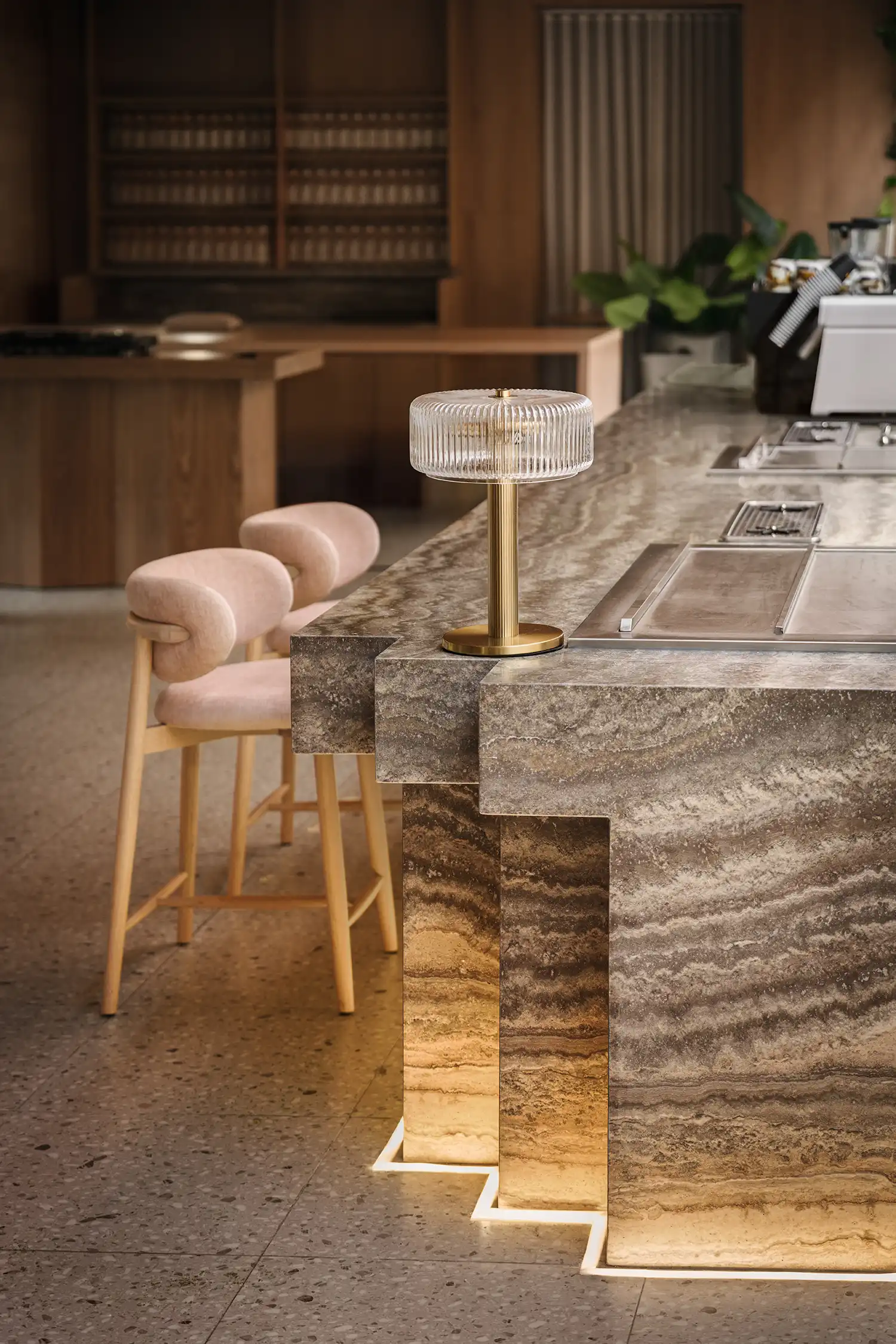 Close-up of a grey travertine bar counter with a fluted glass lamp and pink velvet stools.