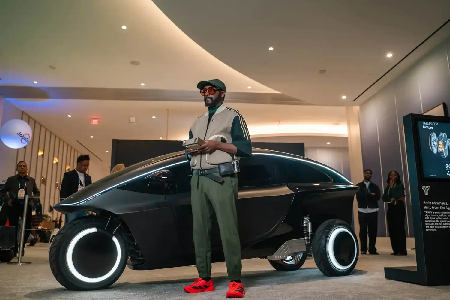 Will.i.am standing next to the Trinity vehicle at an indoor event with a digital screen in the background.
