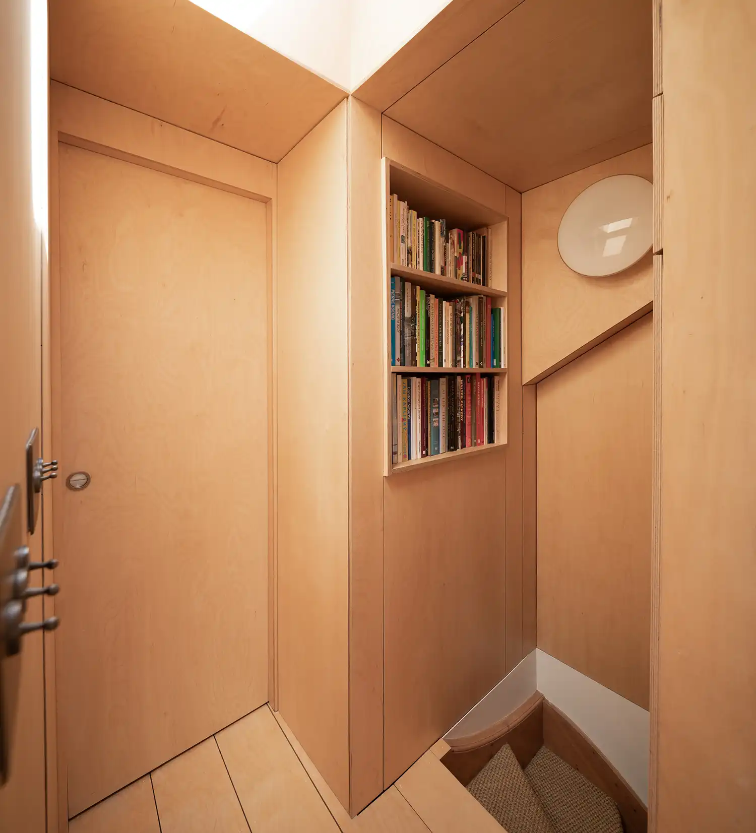 Close-up of a birch plywood doorway and recessed bookshelf in a narrow attic corridor.