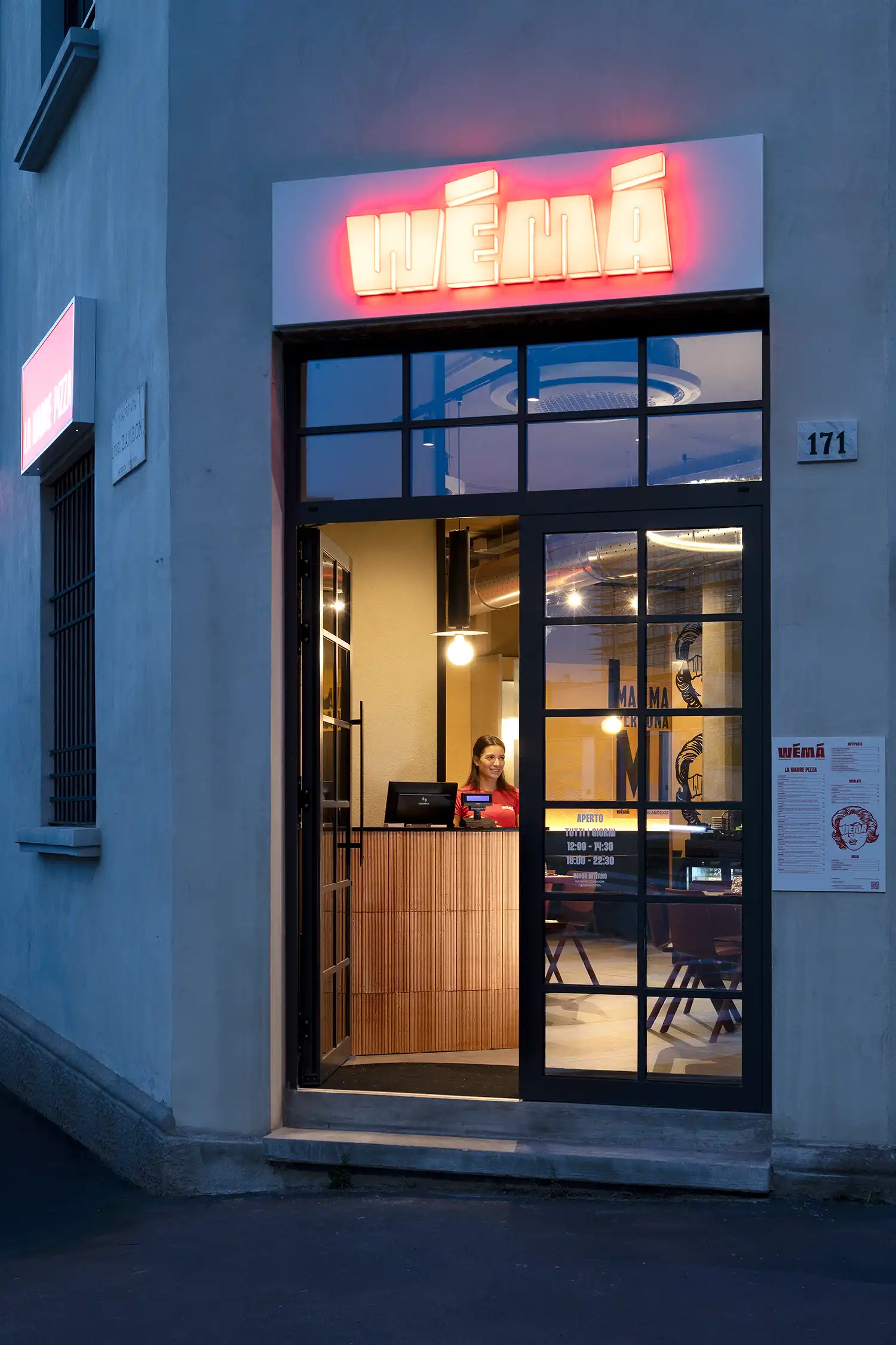 Exterior twilight view of WÉMA pizzeria in Milan Certosa District featuring a red neon sign and industrial window frames.