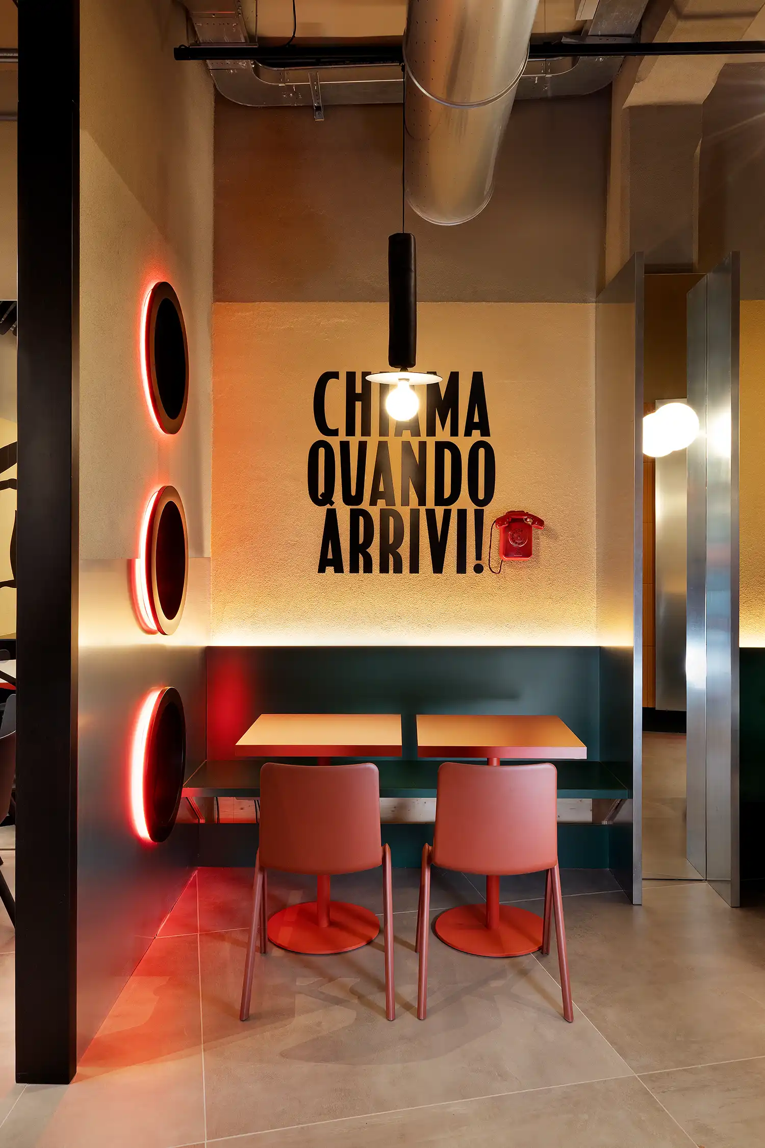 Interior dining nook at WÉMA Milan with "CHIAMA QUANDO ARRIVI!" wall text and red porthole lighting.
