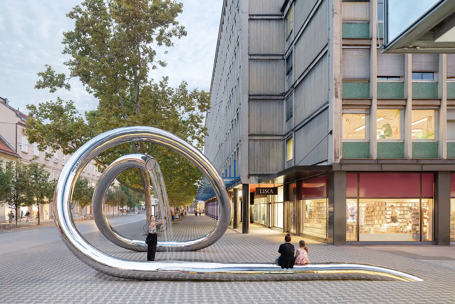 Reflective steel sculpture at dusk with warm light from nearby shop windows.